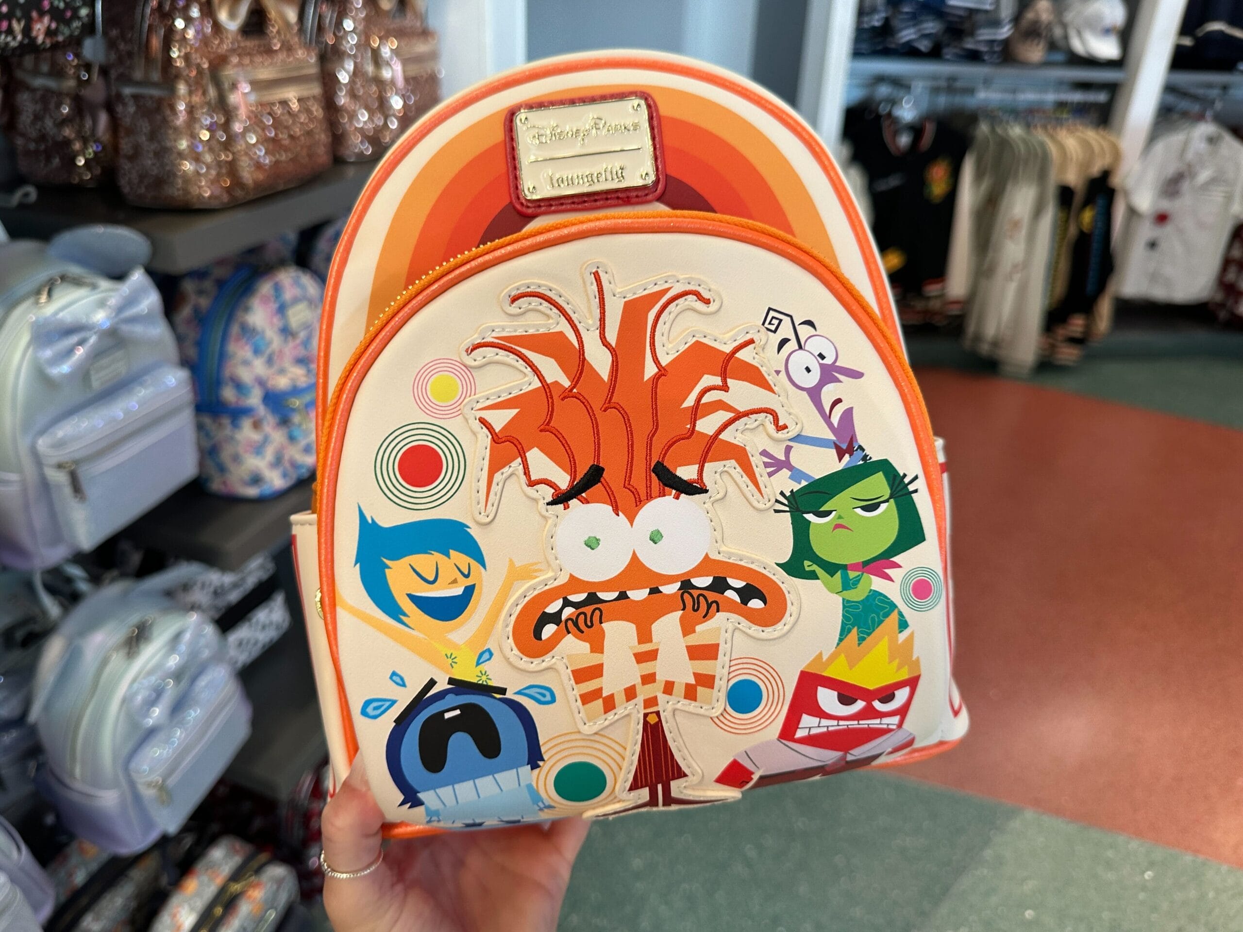 A hand displays an Inside Out 2 Loungefly Mini Backpack with film characters, spotted in a Disney Parks merchandise store display.