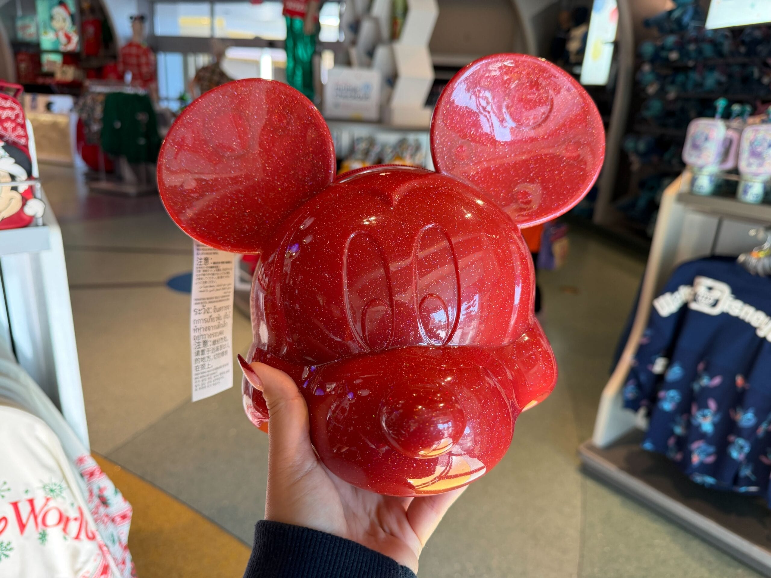 A hand holds a shiny, red Mickey Mouse-shaped Daily Recap container inside a Disney park store, no castle in view to compare.