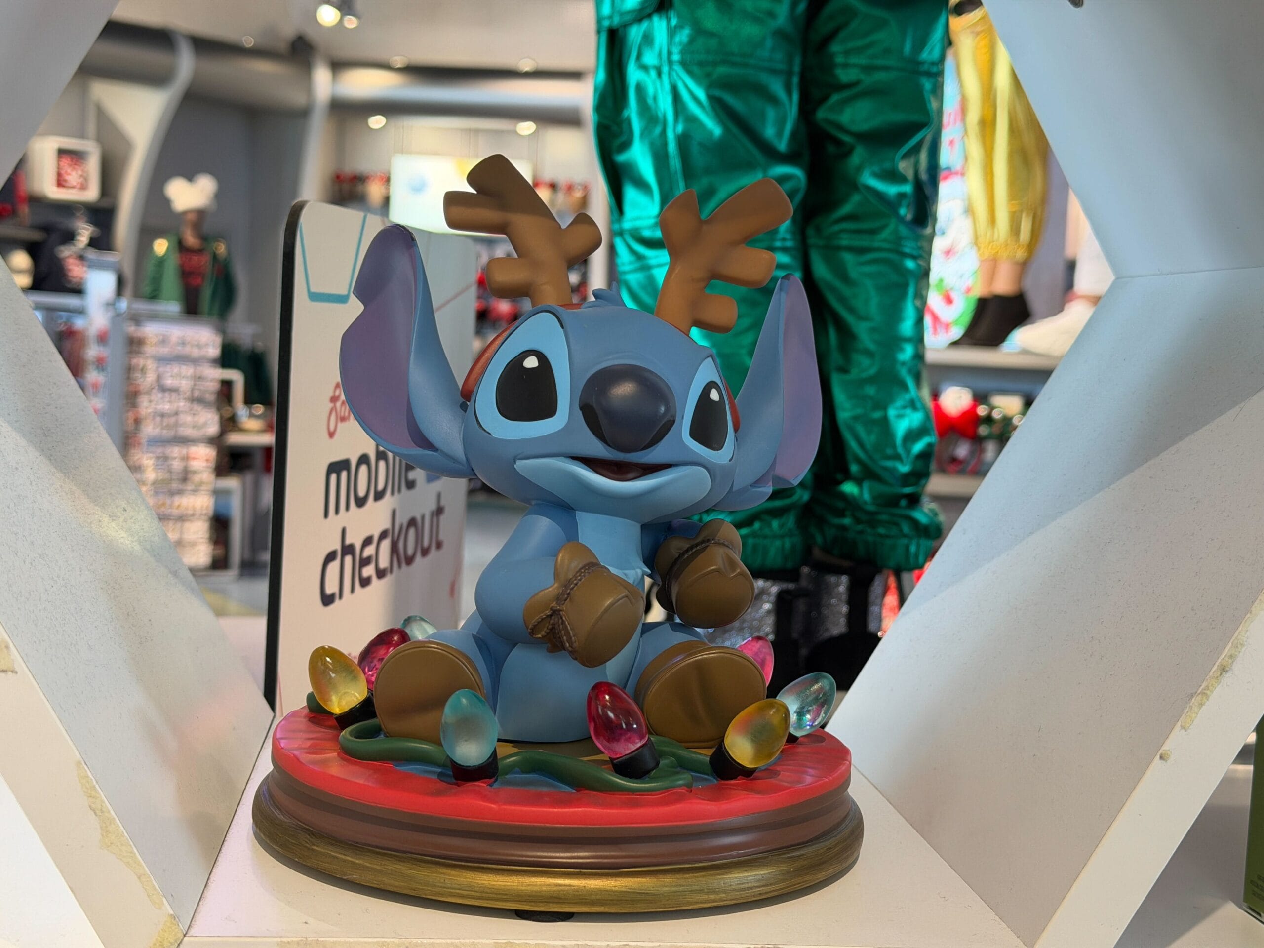 A festive Light-Up Stitch with reindeer antlers sits on a red base in a Disney World holiday display, surrounded by park decor.