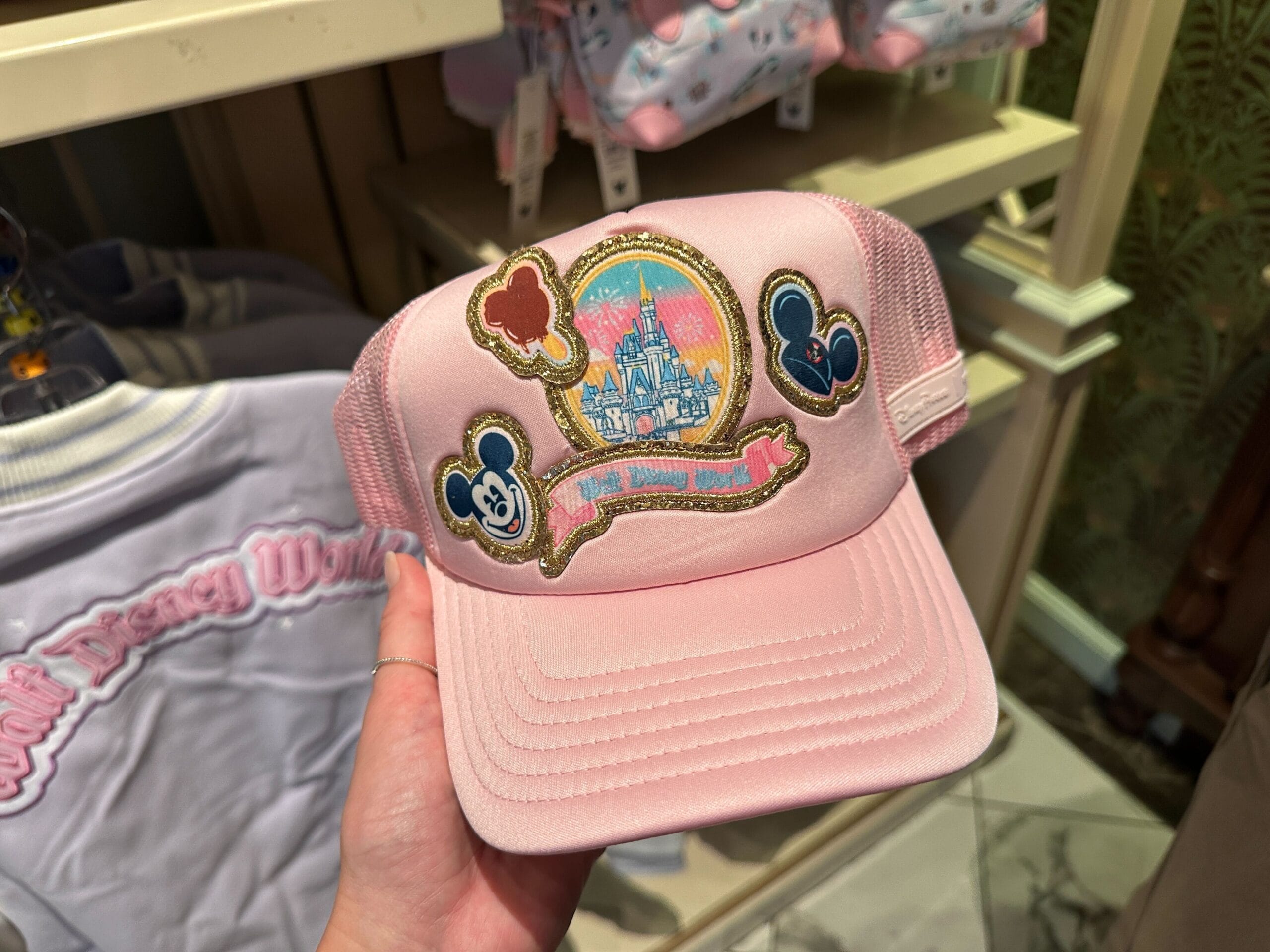 A hand holds a pink Walt Disney World cap featuring Mickey Mouse patches and a castle design resembling Cinderella Castle in Florida.