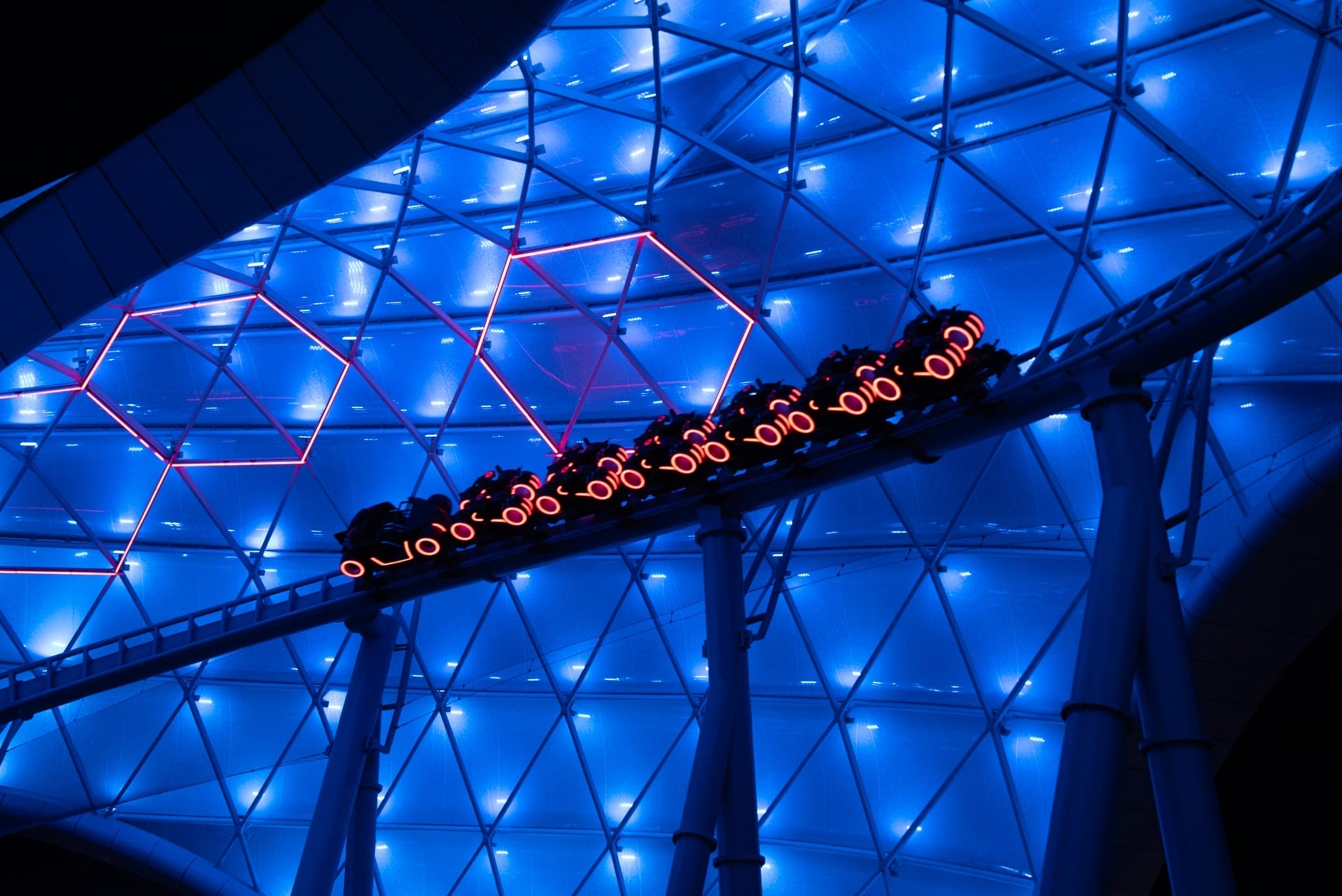 Tron: Ares-inspired Disney ride glowing with blue and pink lights, sleek vehicles racing on a curved night track; no castle in view.