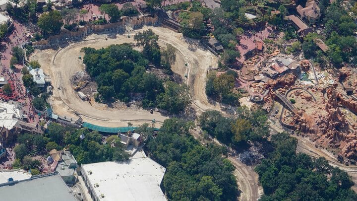 Aerial view of a winding dirt construction site by Tom Sawyer Island, set among lush trees and Disney park attractions—no castle visible.