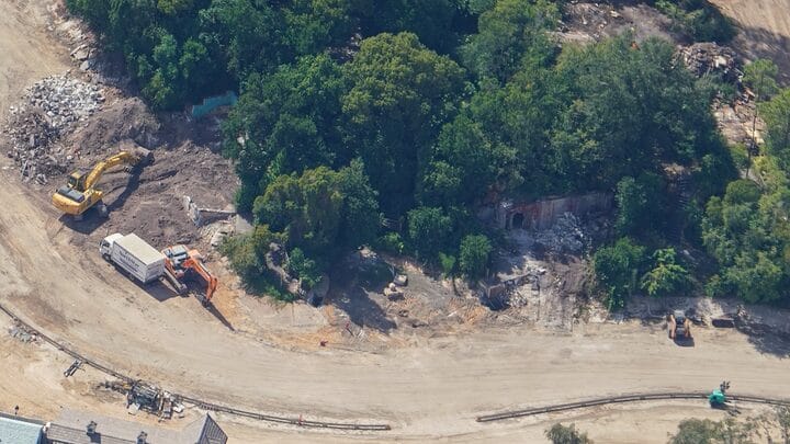 Aerial view of construction vehicles clearing land near a winding dirt road and lush trees, similar to early Disney park expansions.