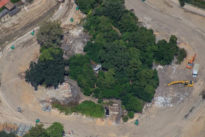 Aerial view of Tom Sawyer Island under construction at a Disney park, ringed by lush trees and work vehicles, no castle visible.