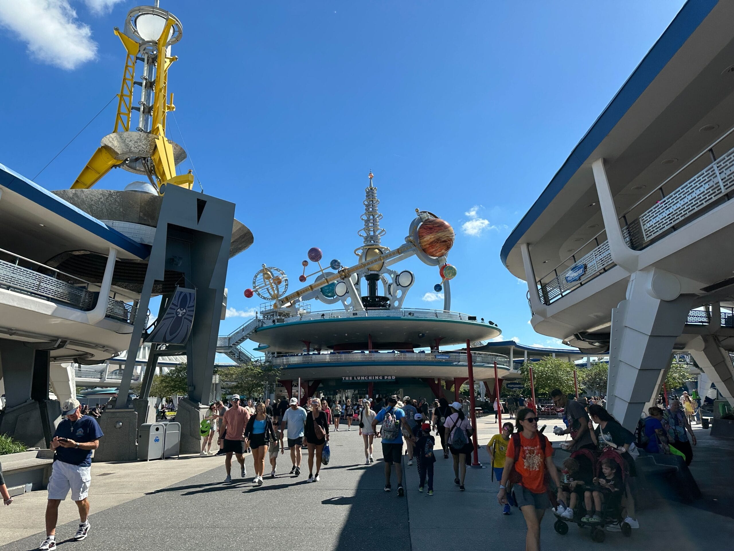 Guests stroll through Tomorrowland at a Disney park, Astro Orbiter closed for refurbishment, beneath a vivid blue sky.