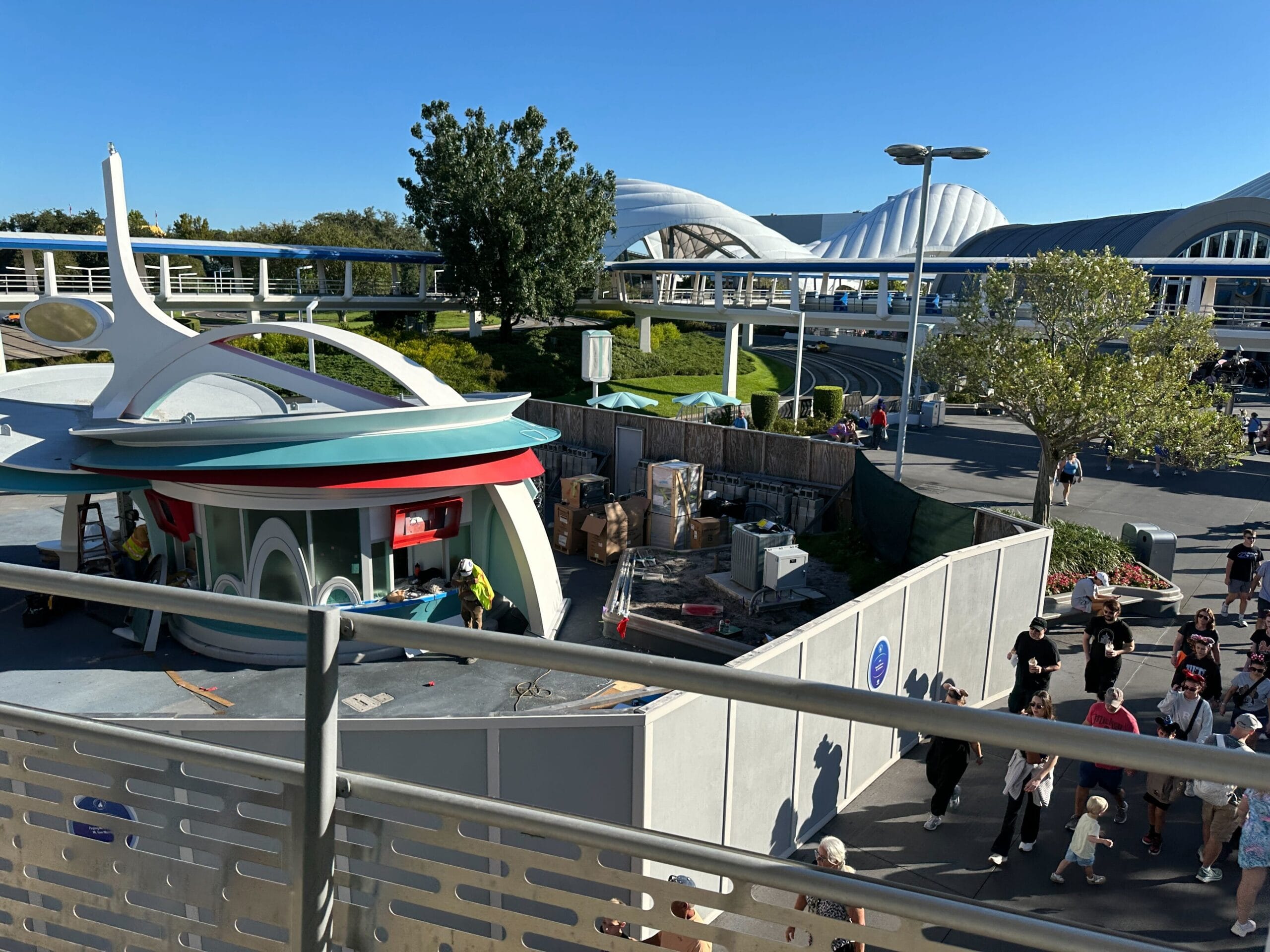 Workers and barriers at a Disney park construction zone, beside menu boards, with crowds and clear sky—no castle in view to compare.