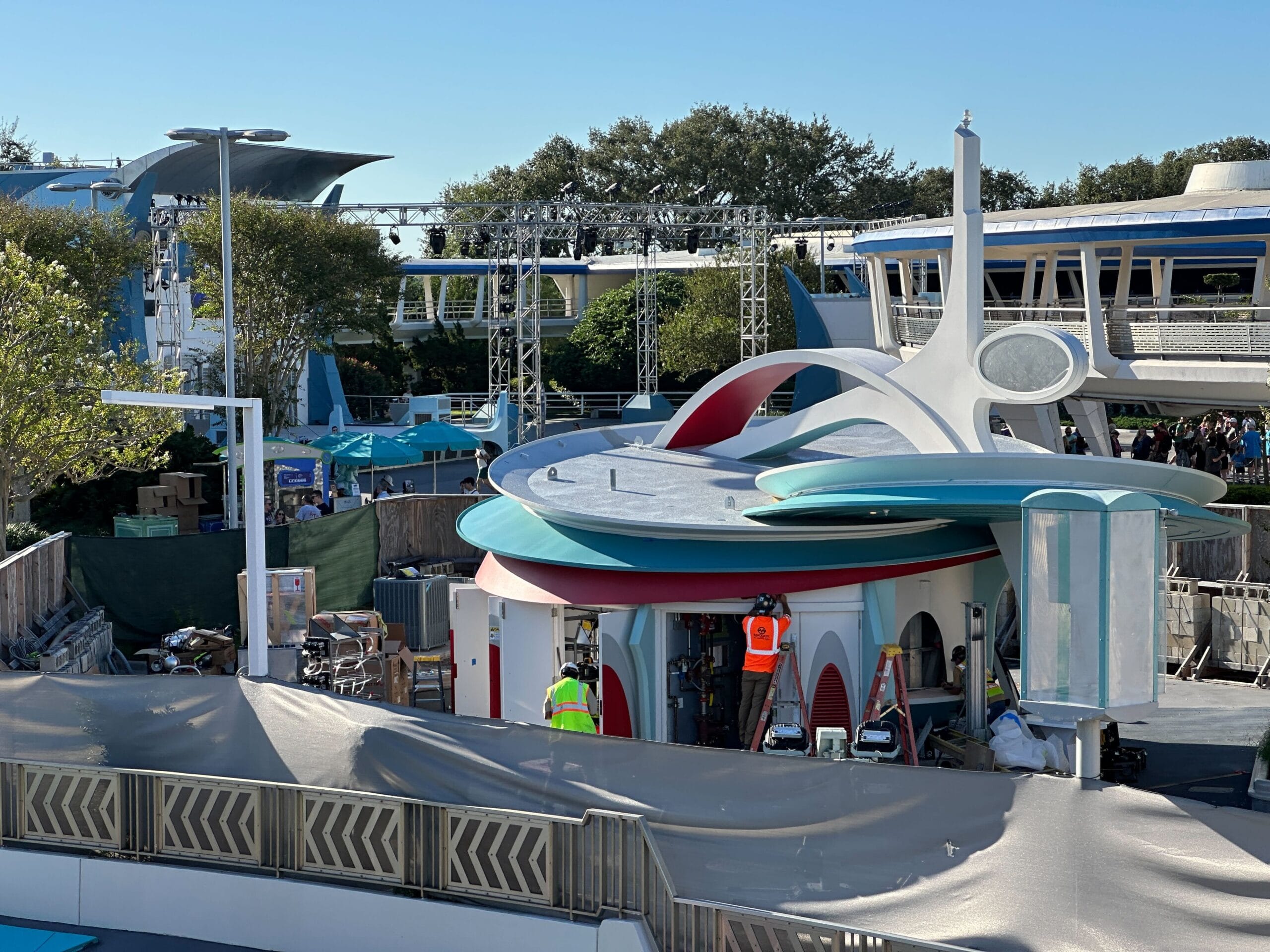 Construction workers in vests build a futuristic round Cool Ship at Disney World, with barriers and gear, no castle in view.