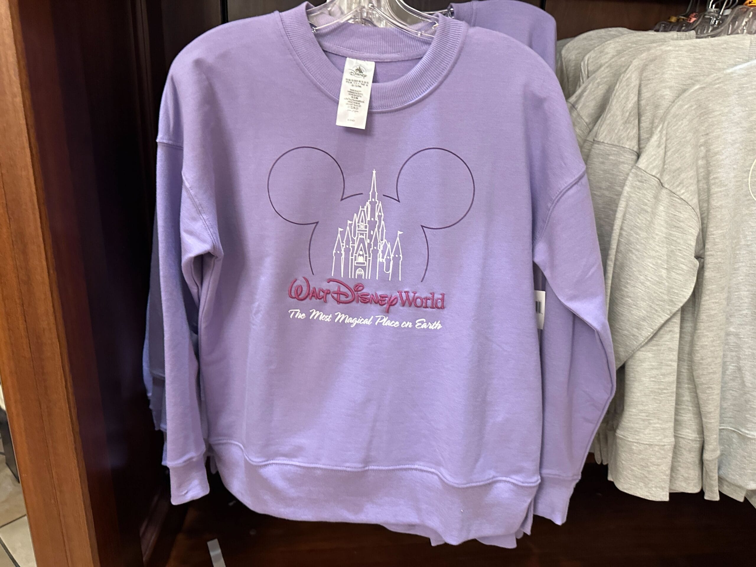 A lavender Walt Disney World sweatshirt featuring a Mickey Mouse outline and a Cinderella Castle graphic, like Orlando's castle, displayed in a store.