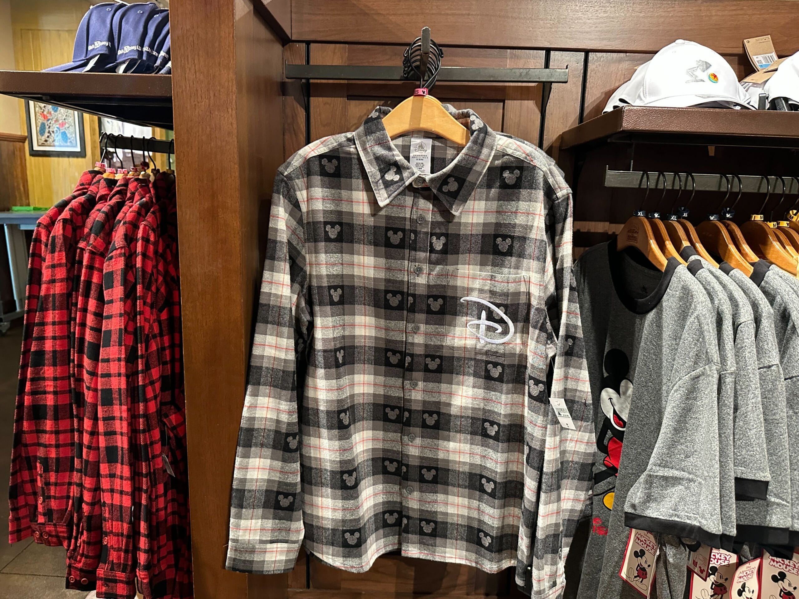 A gray plaid shirt featuring tiny Mickey Mouse heads and a white Disney "D" logo hangs on a wooden rack in a Disney Park store.