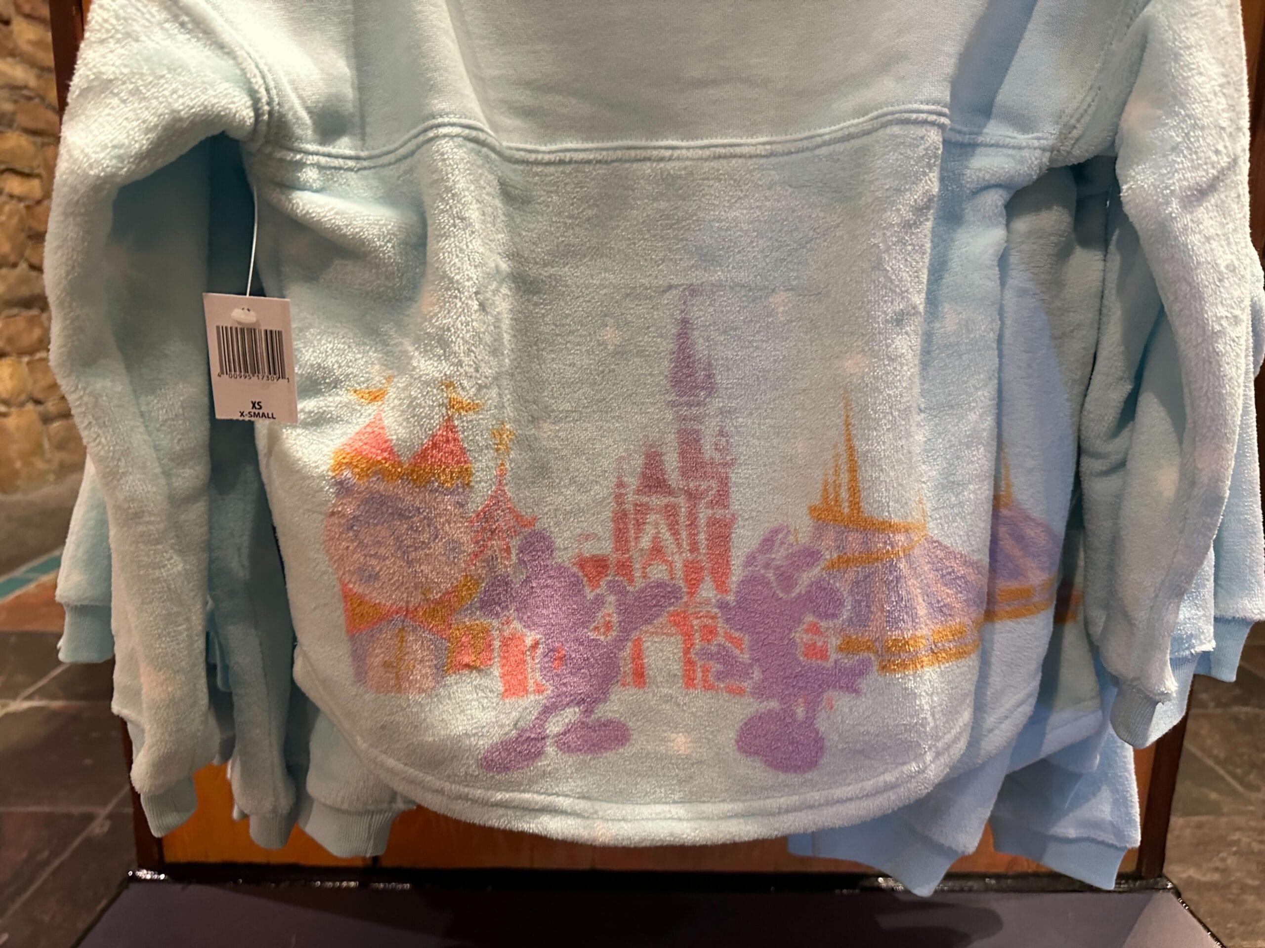 Light blue fleece jacket features pastel Mickey, Minnie, and a Disney castle similar to Cinderella Castle at Magic Kingdom on display.