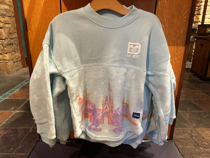 Light blue children’s sweater featuring Figment and a Disney castle resembling Cinderella Castle from Walt Disney World, on a store rack.