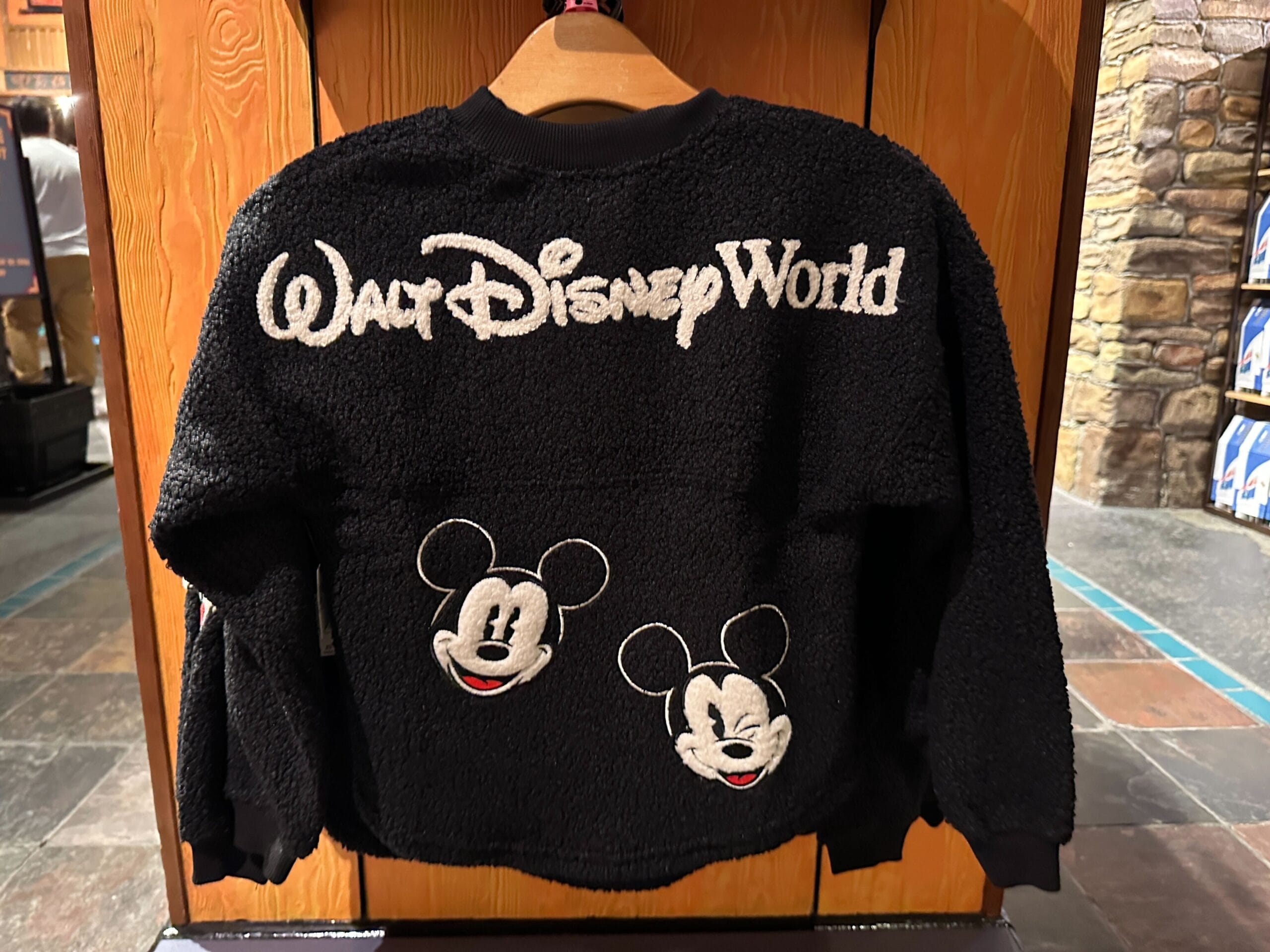 A black fuzzy Walt Disney World sweater with two Mickey Mouse faces on the back, hanging on a wooden display in a park shop.