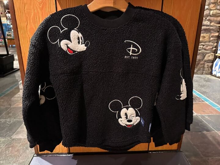 Black fleece sweater with embroidered Mickey Mouse faces and Disney logo, hanging on a wooden hanger, ideal for Disney park visits.