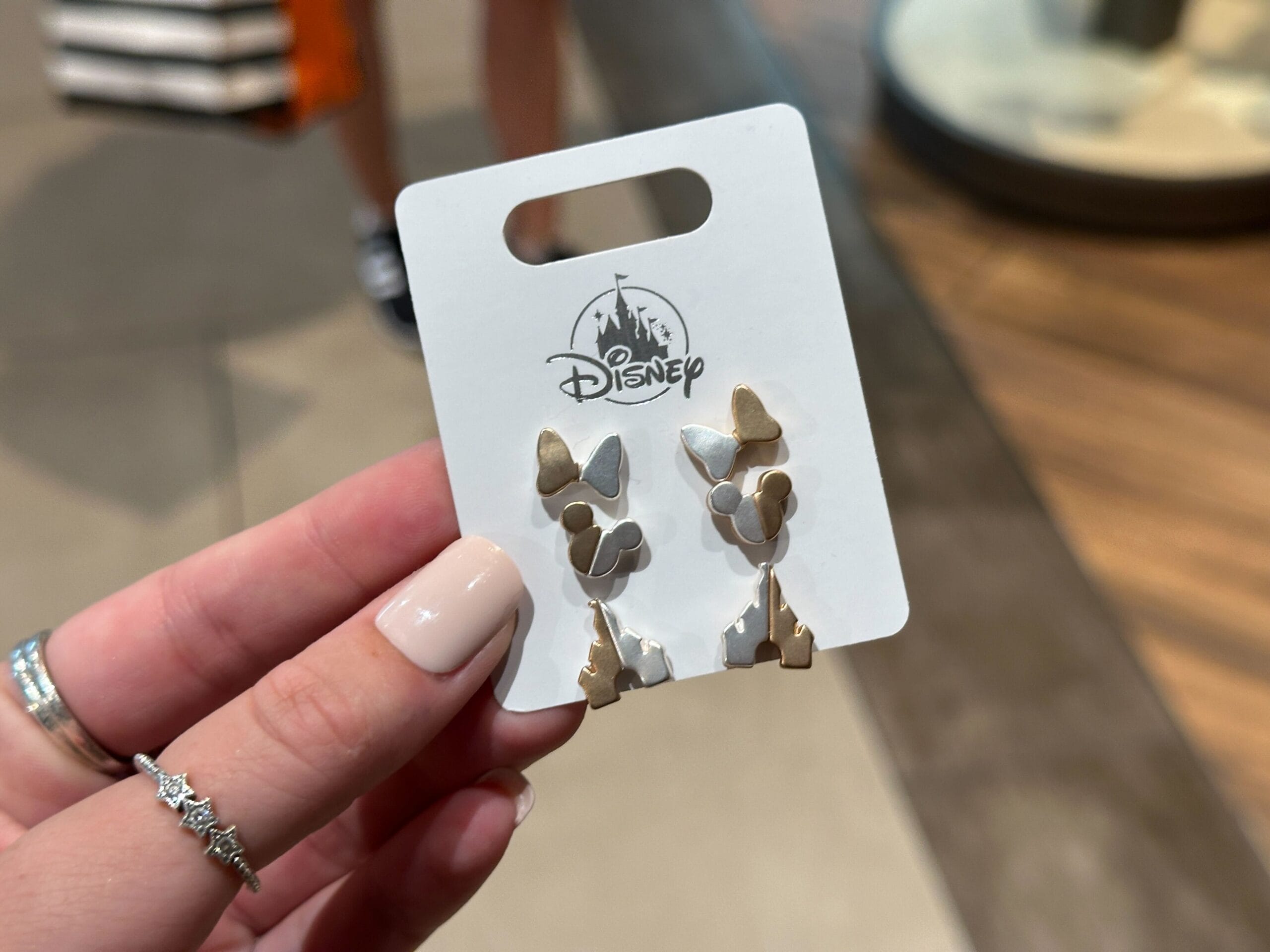 A hand holds a Disney Parks card showing silver Minnie and Mickey Mouse silhouette earrings, reminiscent of classic park souvenirs.