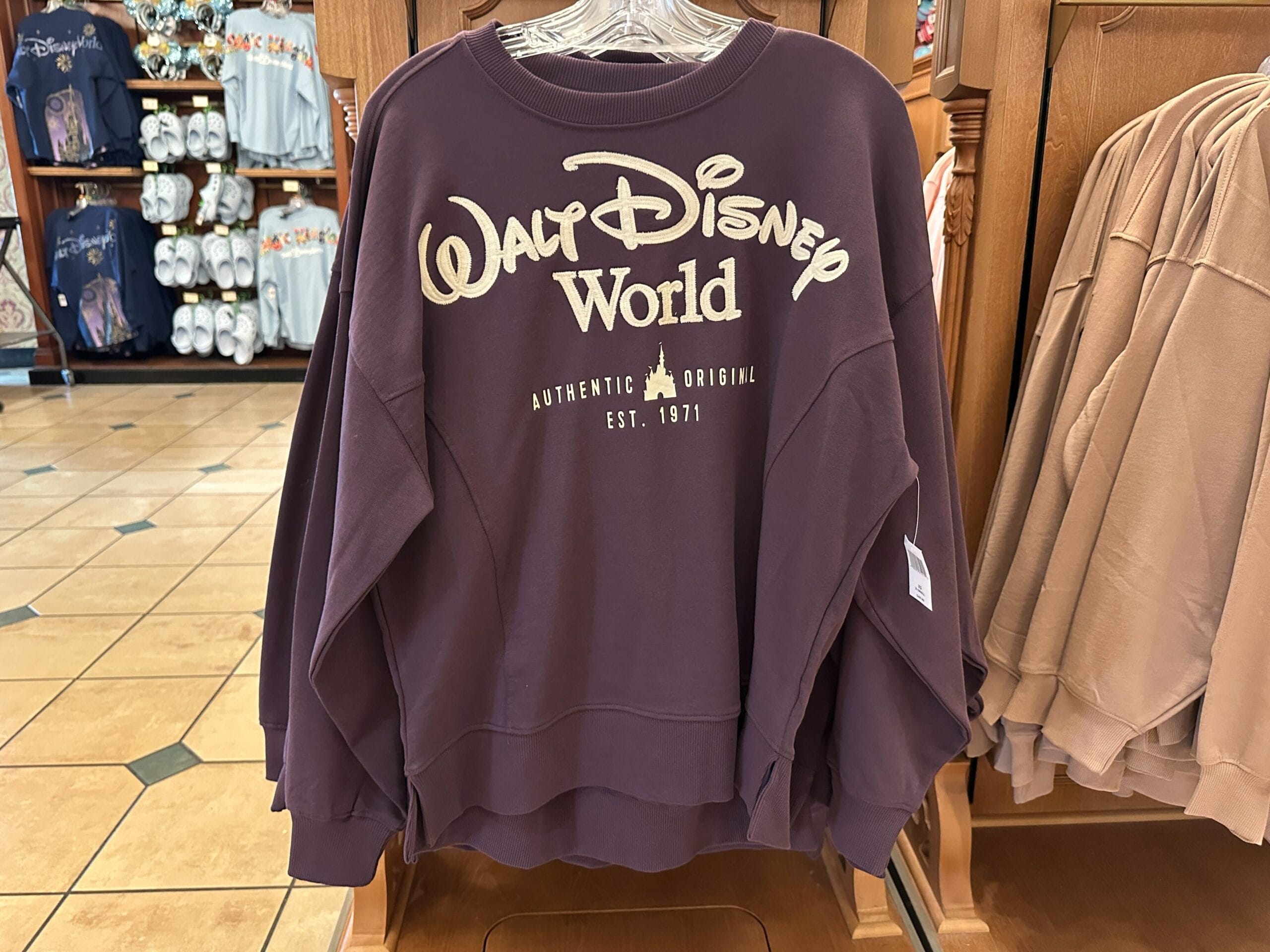 A maroon Walt Disney World sweatshirt with cream lettering hangs on a rack in a park gift shop, unique to the Florida resort.