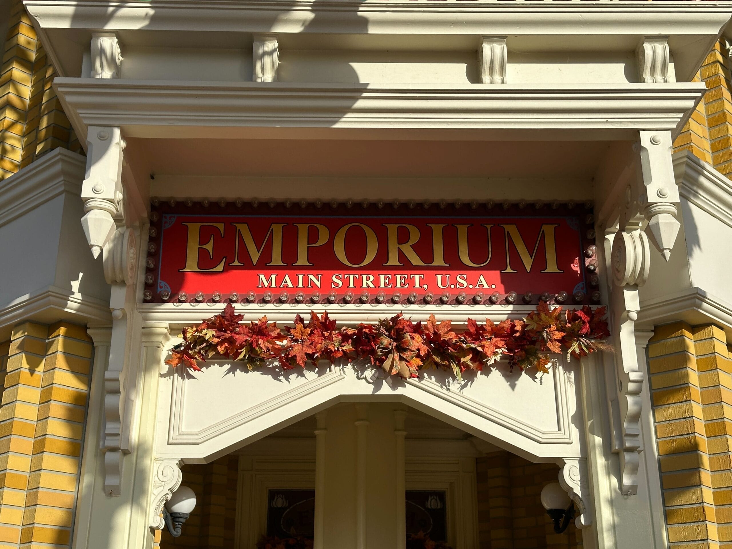 A red "EMPORIUM MAIN STREET, U.S.A." sign hangs above a doorway adorned with autumn leaves, echoing classic Disney park style.