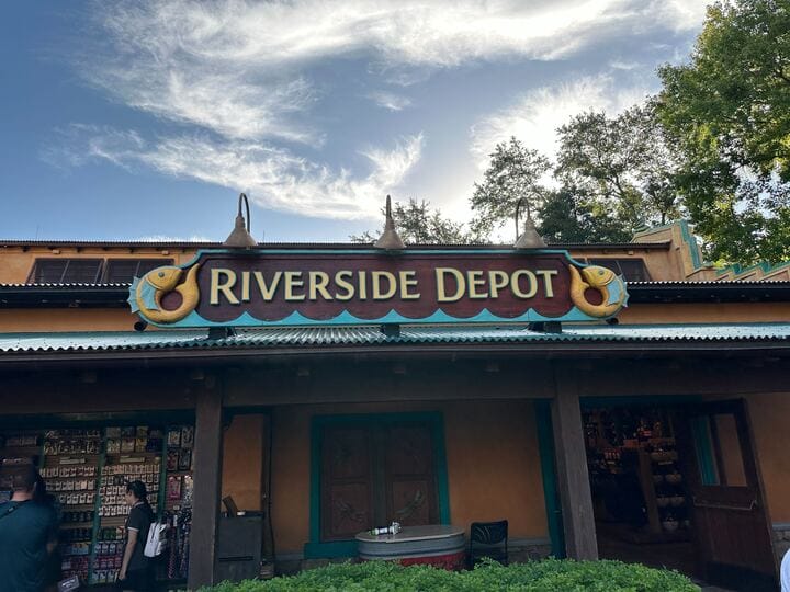 Riverside Depot shop sign at Disney park, surrounded by themed lamps and lush trees beneath a bright, cloud-dotted blue sky.