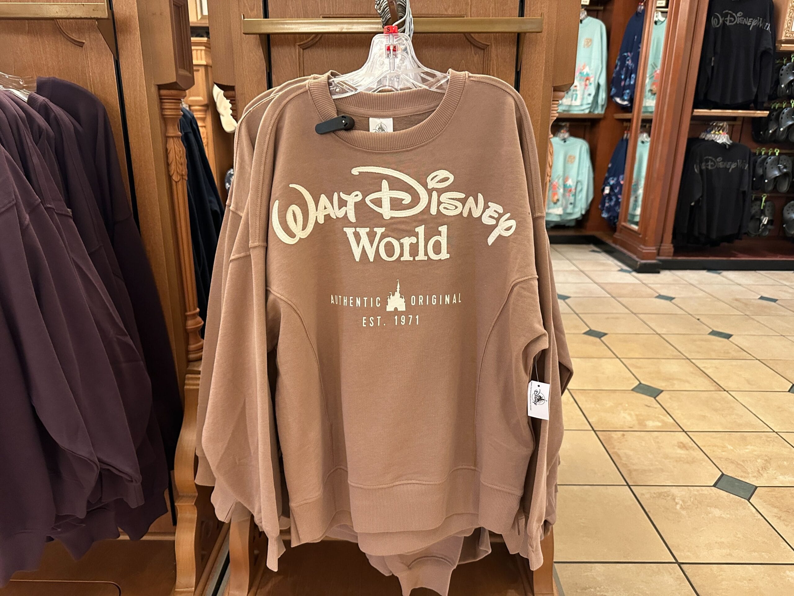 A brown "Walt Disney World" sweatshirt hangs in a shop, its design evoking classic park style, wooden shelves and tile floor.