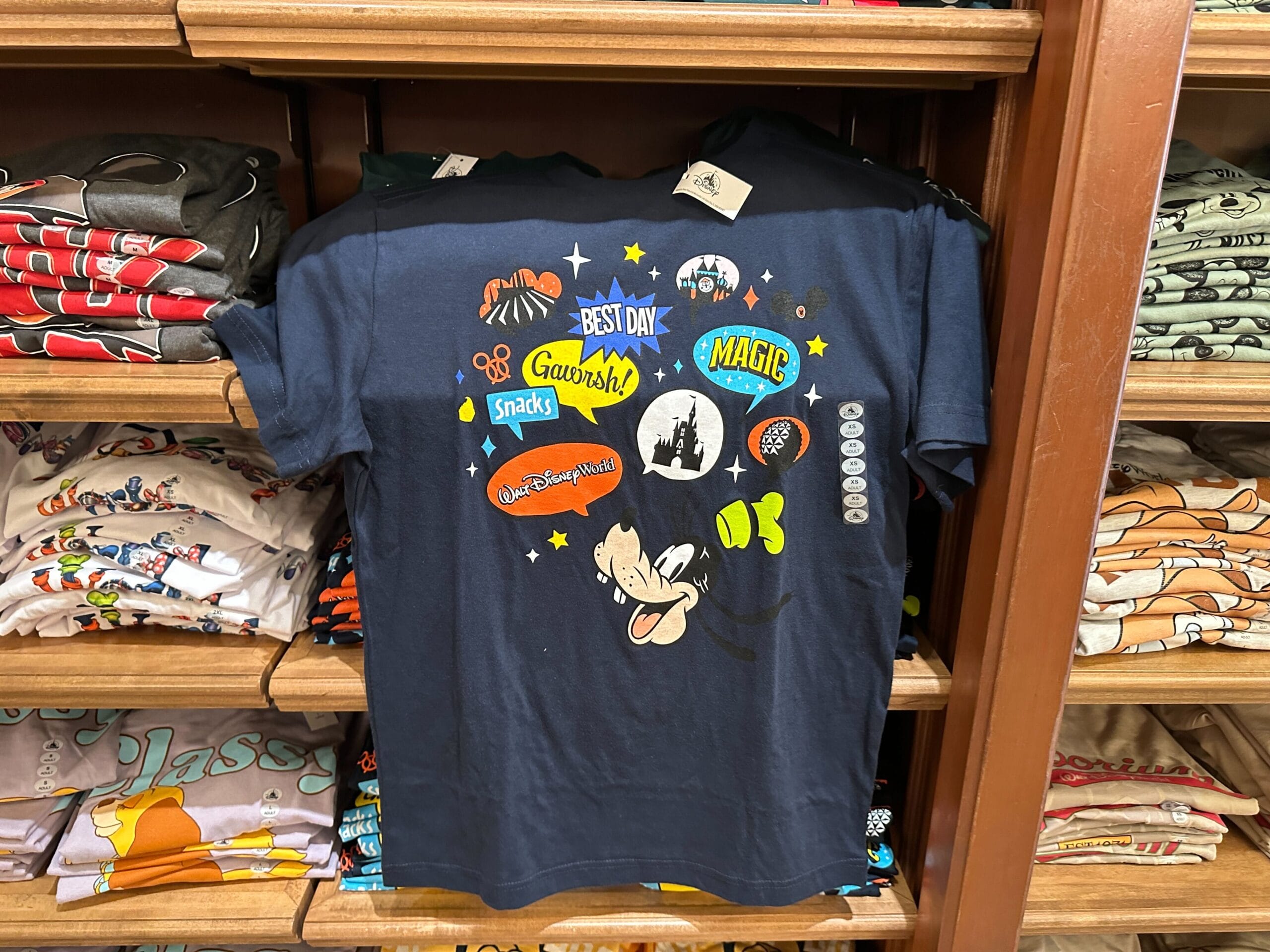 Navy blue Disney Parks t-shirt with themed graphics and words, folded on a shelf with other shirts, reminiscent of park merchandise.