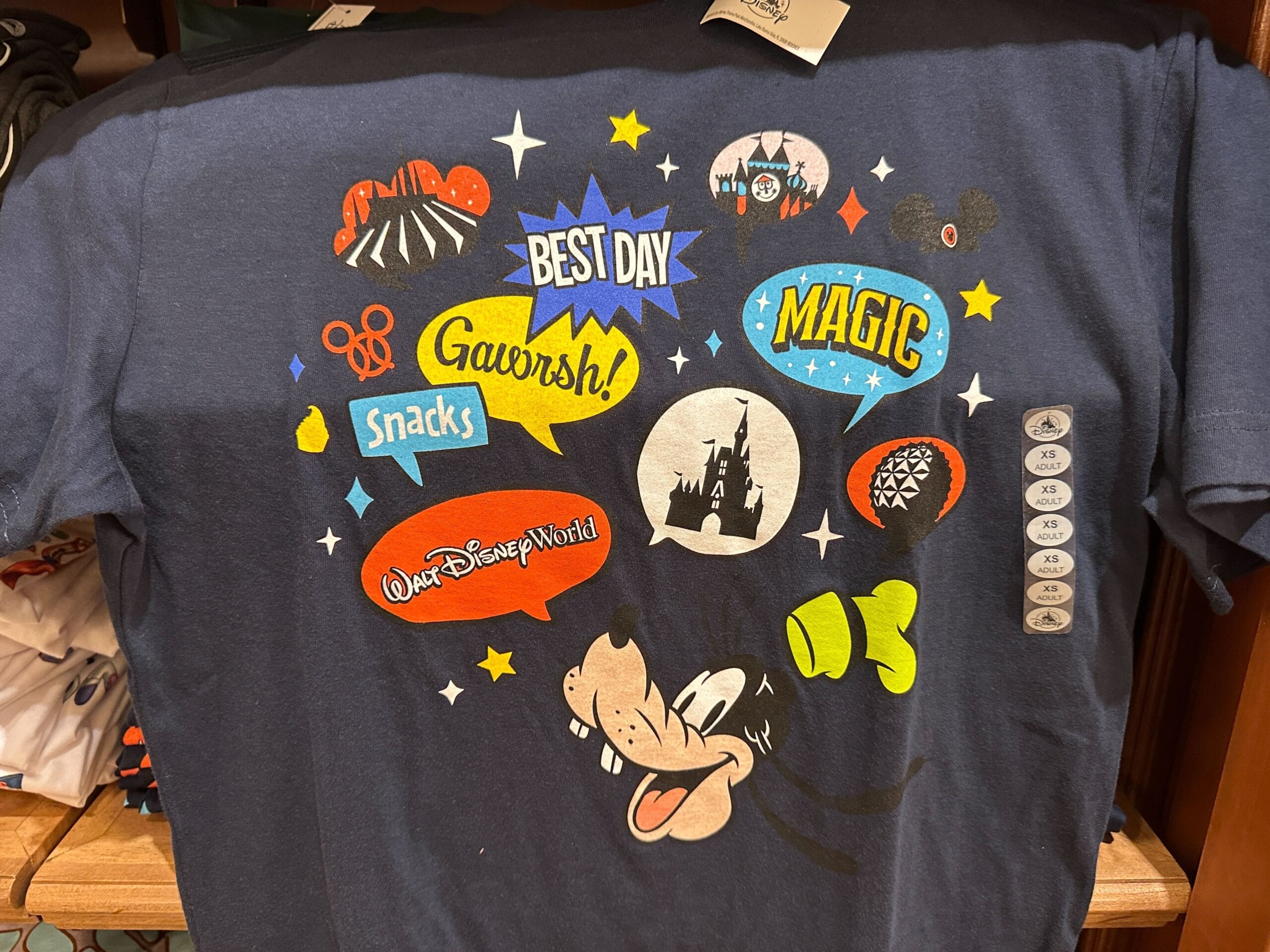 Navy Disney Parks shirt with Goofy, colorful speech bubbles like "Best Day" and "Magic," and park icons, no castle pictured.