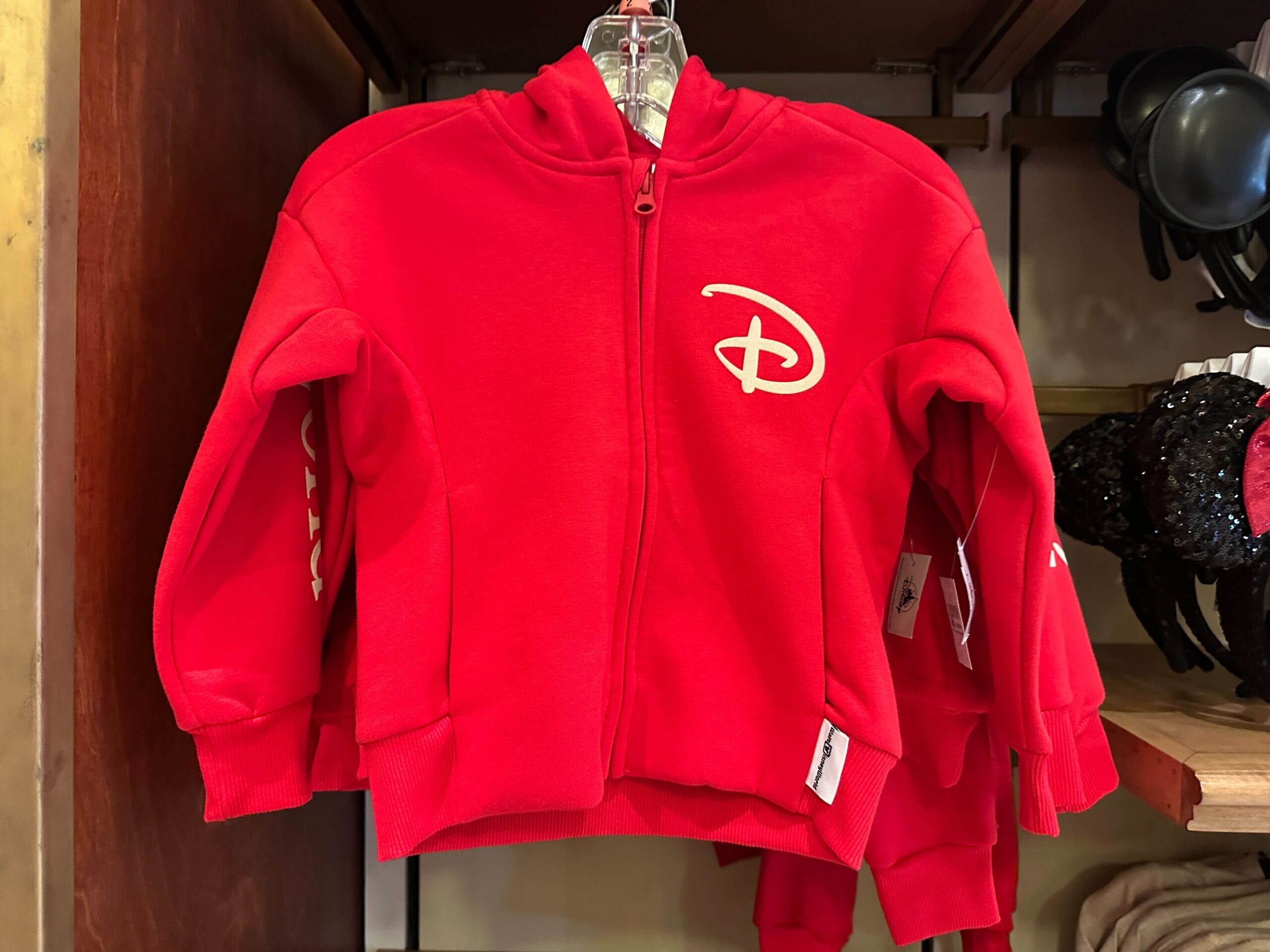 Red kids' zip-up hoodie with a classic Disney-style "D" logo, hanging on a store rack in a Walt Disney Park shop.