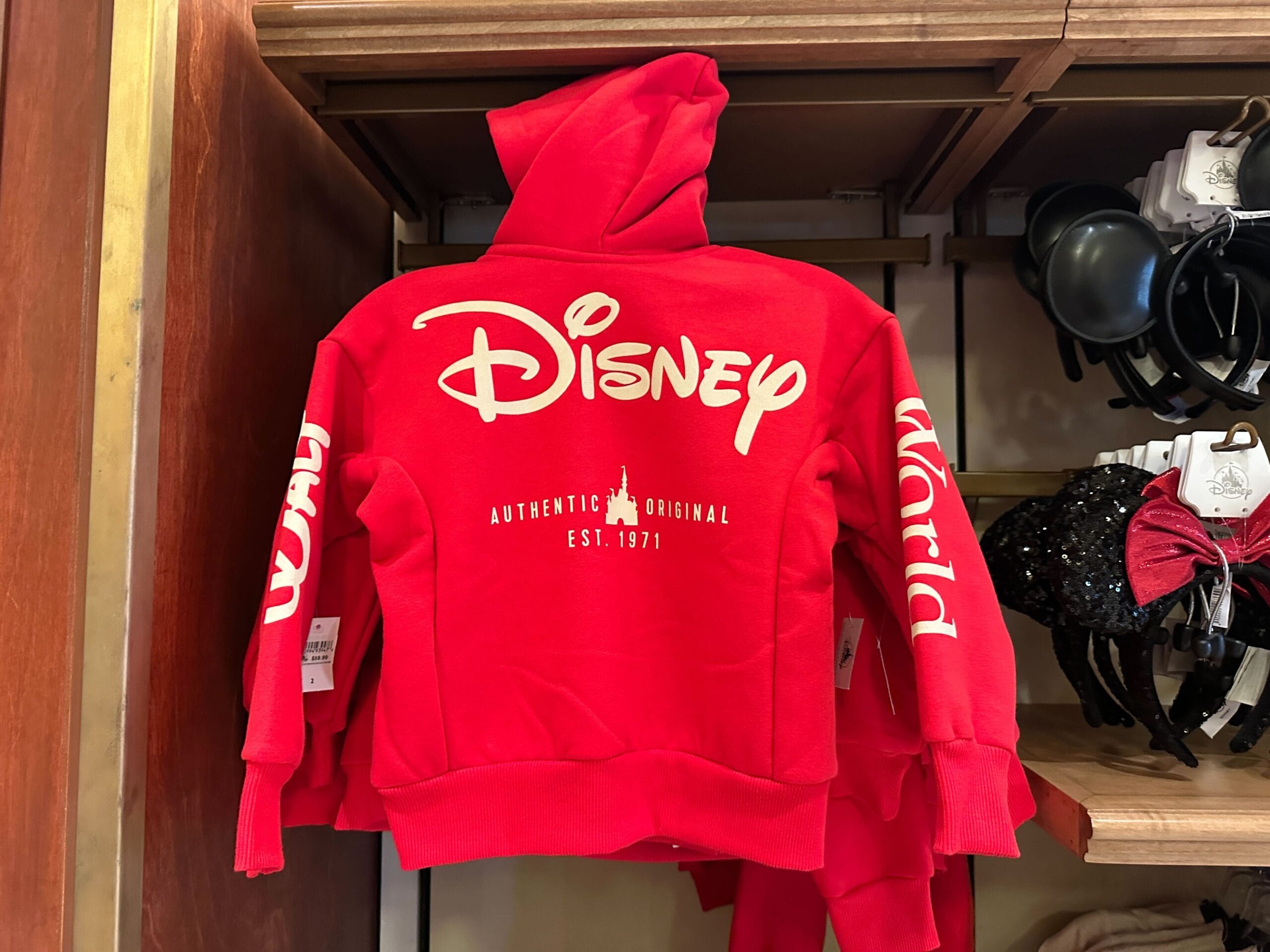 Red Disney Parks hoodie with white lettering, "Disney" logo and "World" on the sleeves, seen on a park shop shelf.