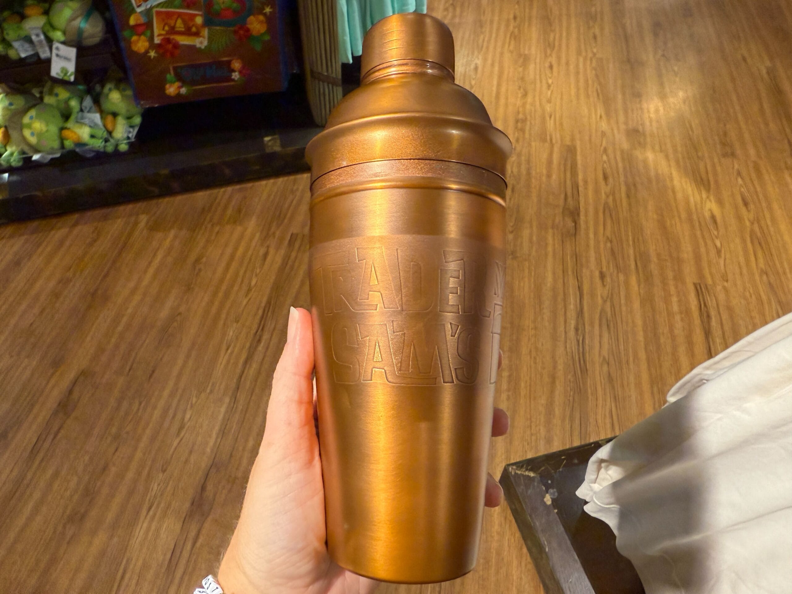 A hand holds a metallic bronze Trader Sam's Shaker, engraved with text, reminiscent of Disney tiki bars, on a wooden floor.