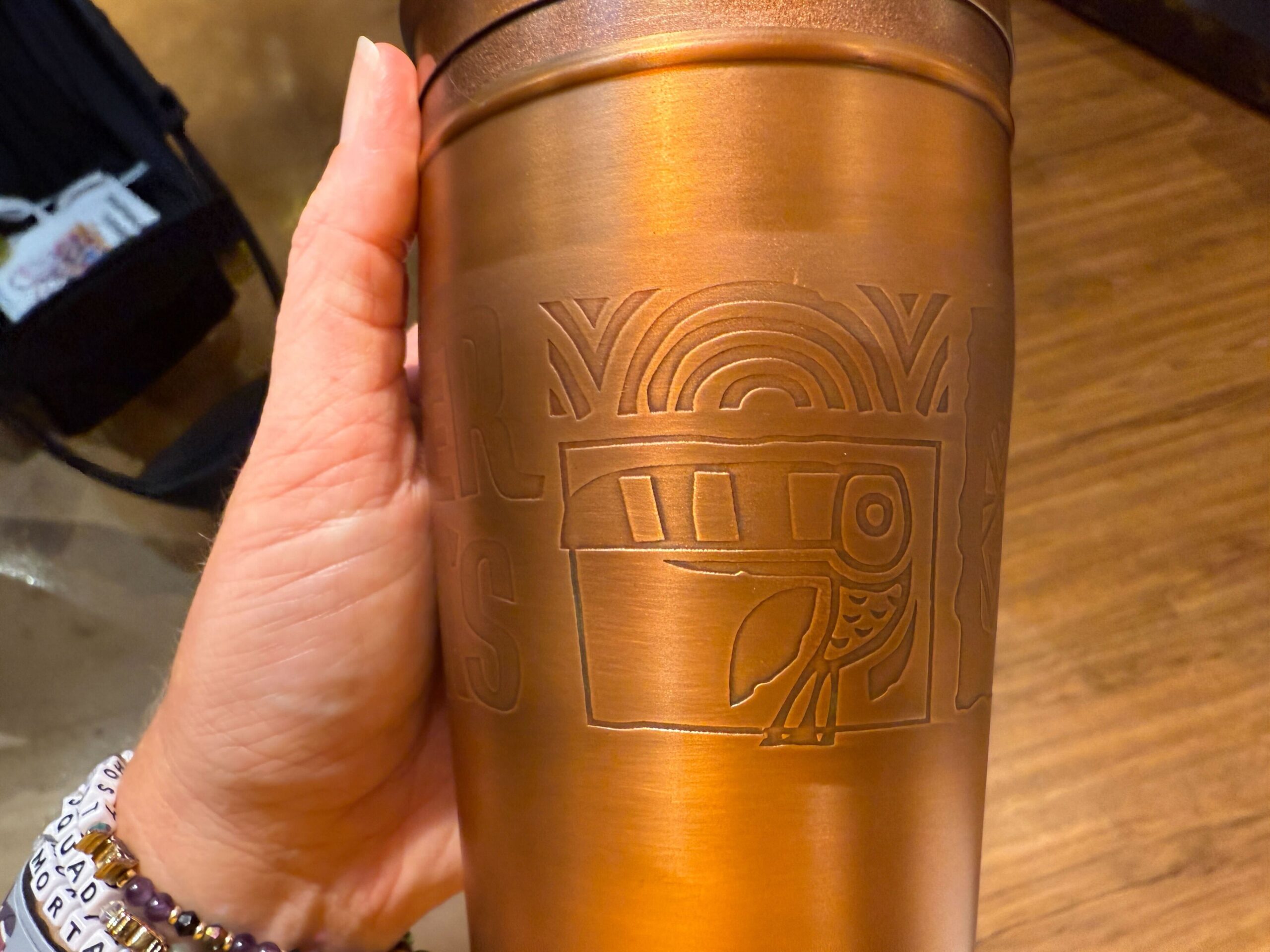 A hand holds a bronze tumbler with engraved rainbow and bird, evoking Trader Sam's cocktail vibes at a Disney Park bar.