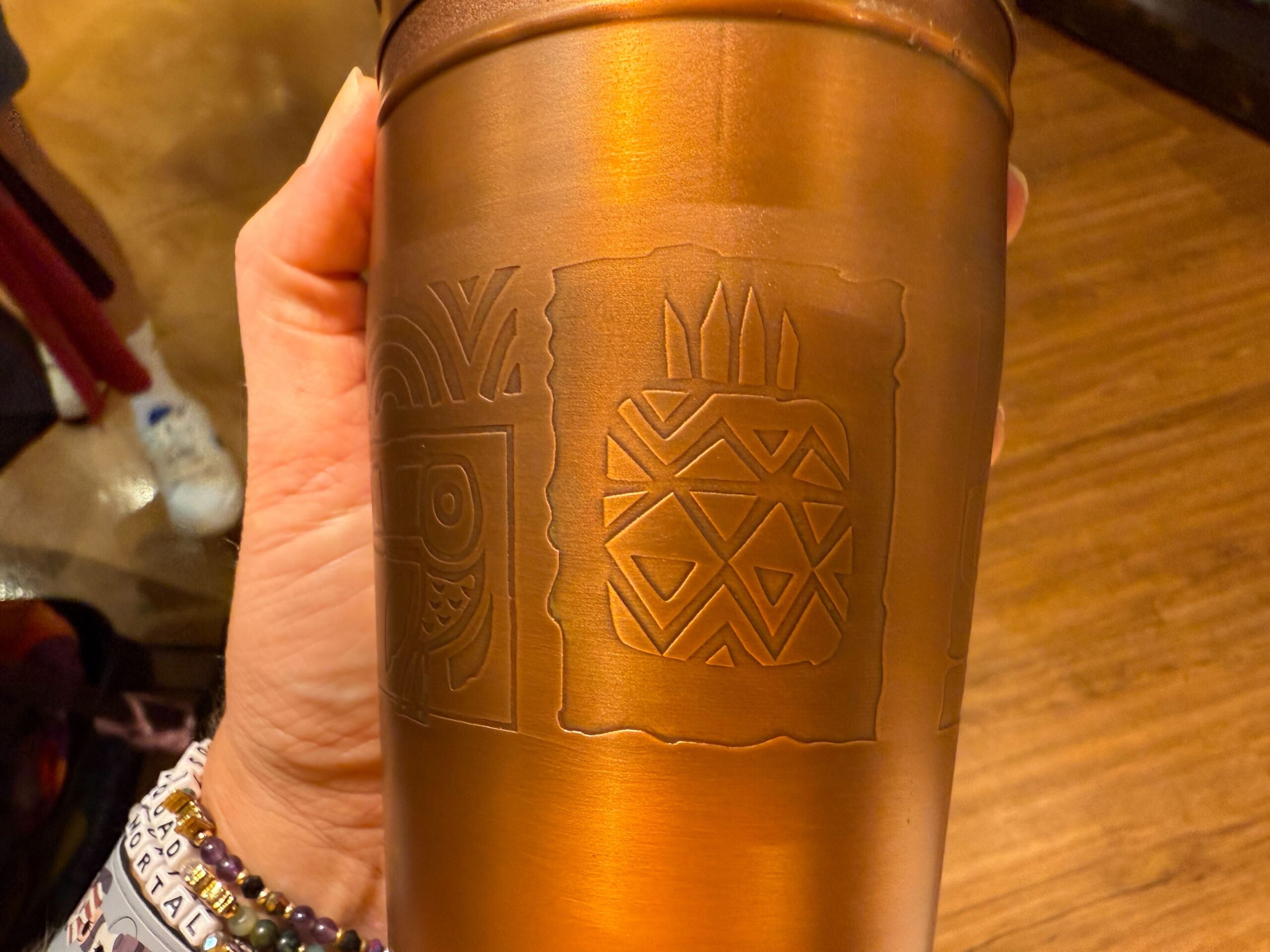 A hand holds a bronze Trader Sam's Shaker cup with geometric, tribal designs—one of the signature souvenirs at Disney parks.