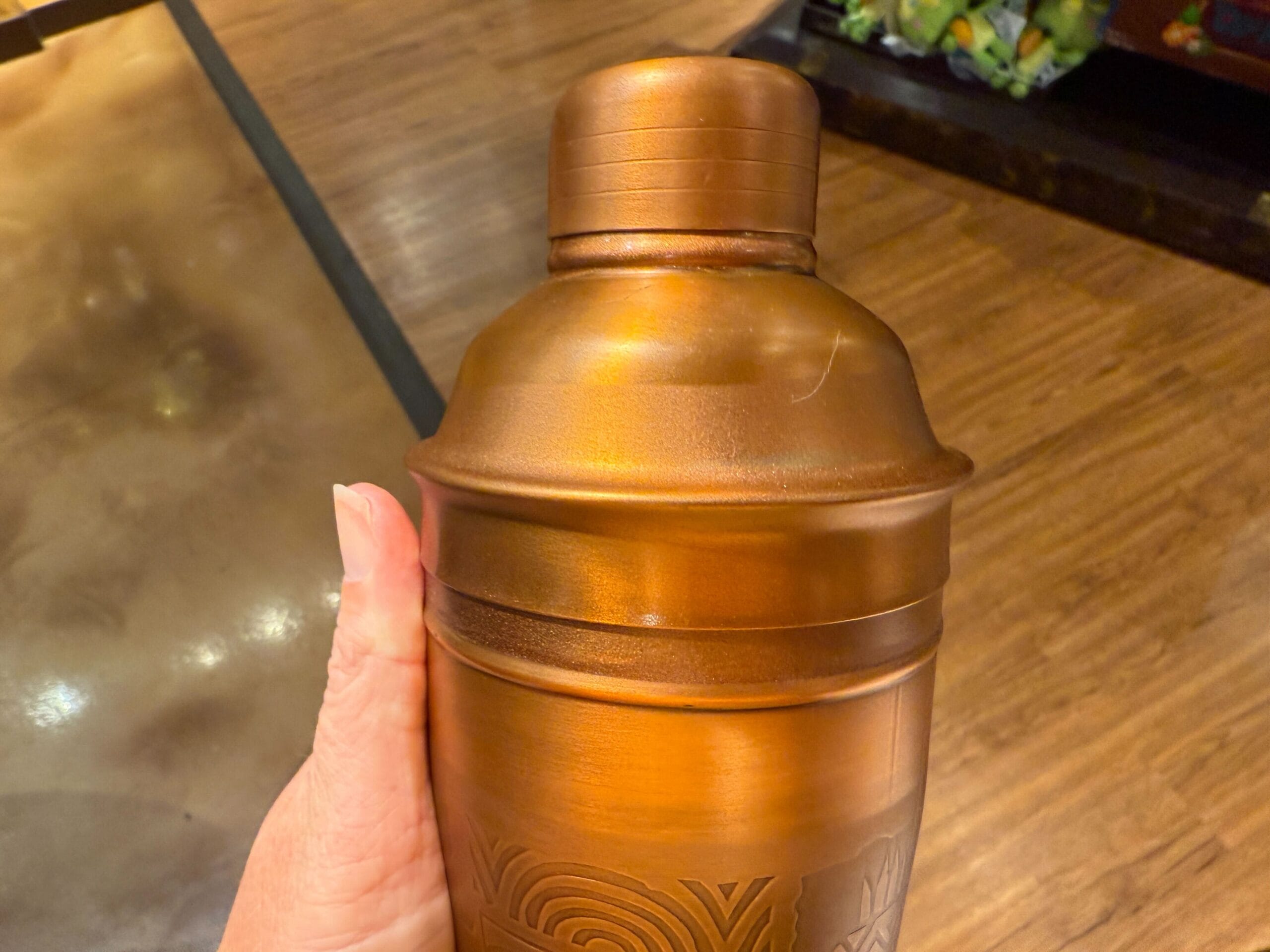 A hand holds a Trader Sam's Shaker for cocktails, reminiscent of Disney's tropical bars, against a wooden floor background.