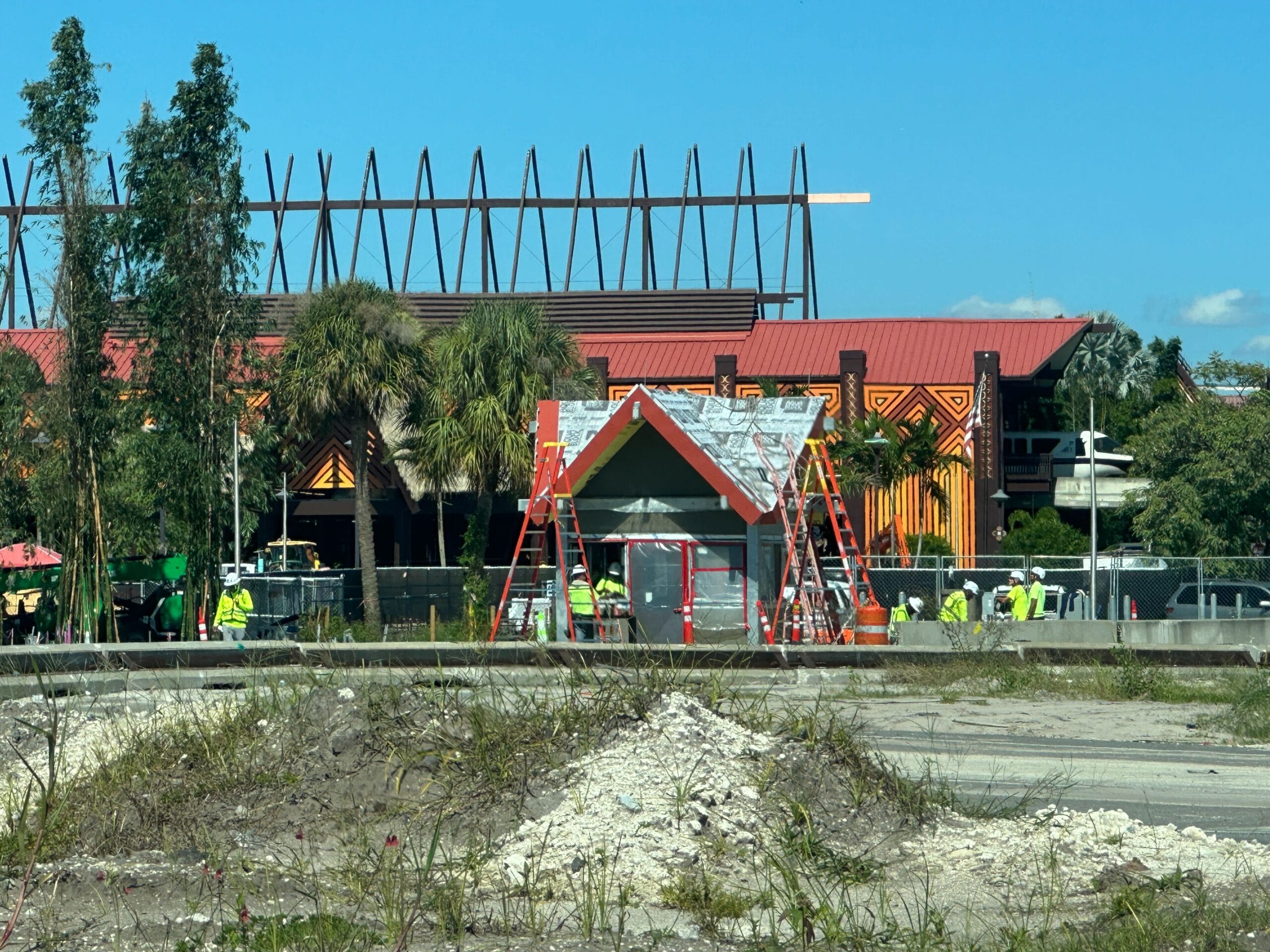 A Polynesian-style guard shack under construction at a Disney park, with workers, ladders, and doors and windows being installed.