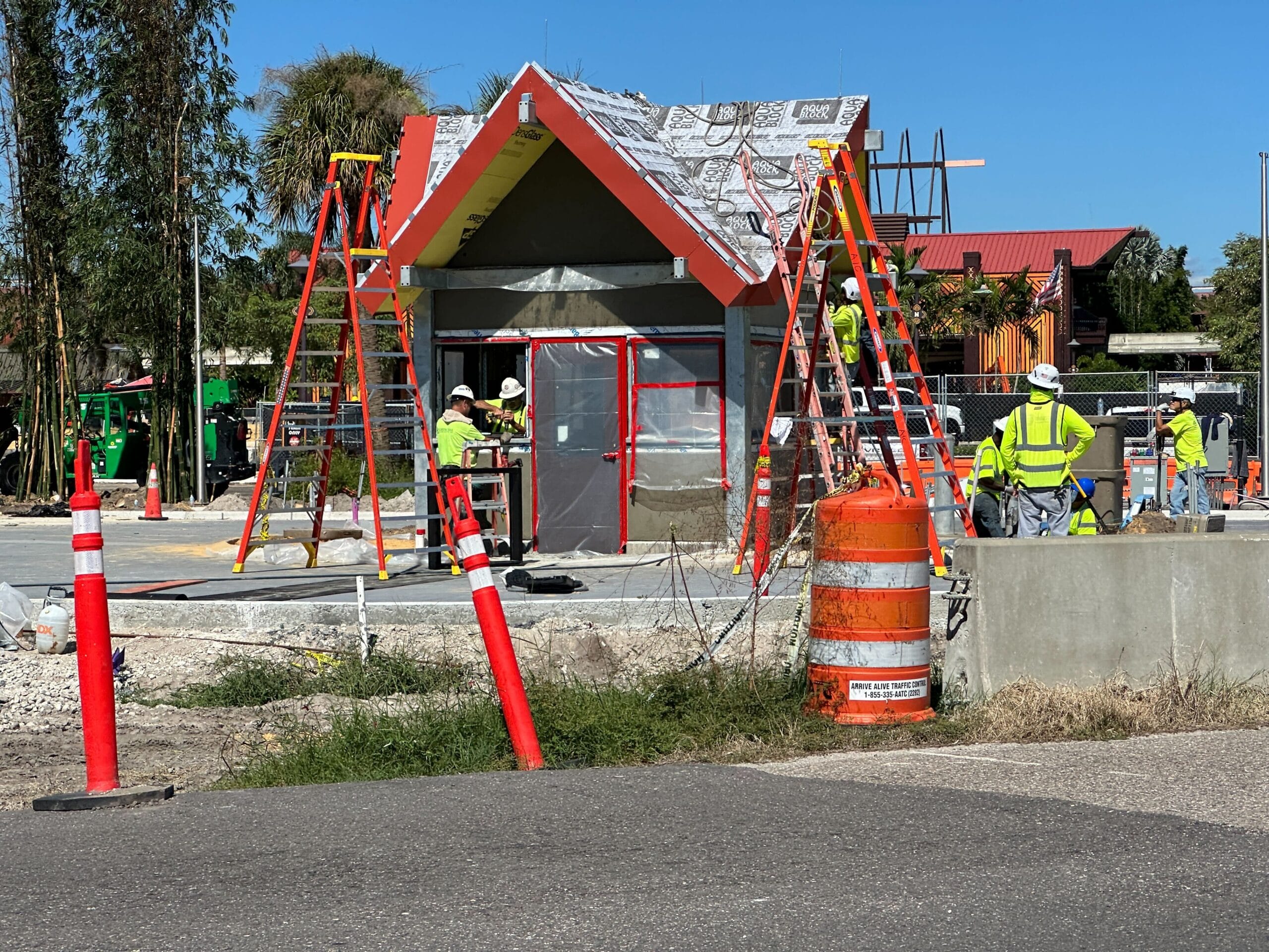 Construction workers in safety gear build a Polynesian Guard Shack at a Disney park, focused on door installation and finishing touches.
