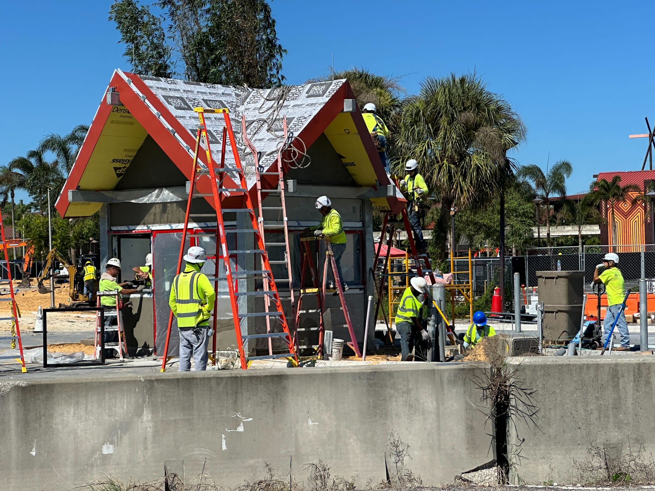 Construction workers in safety vests build a Polynesian-style guard shack, similar to Disney resort theming, under a clear blue sky.