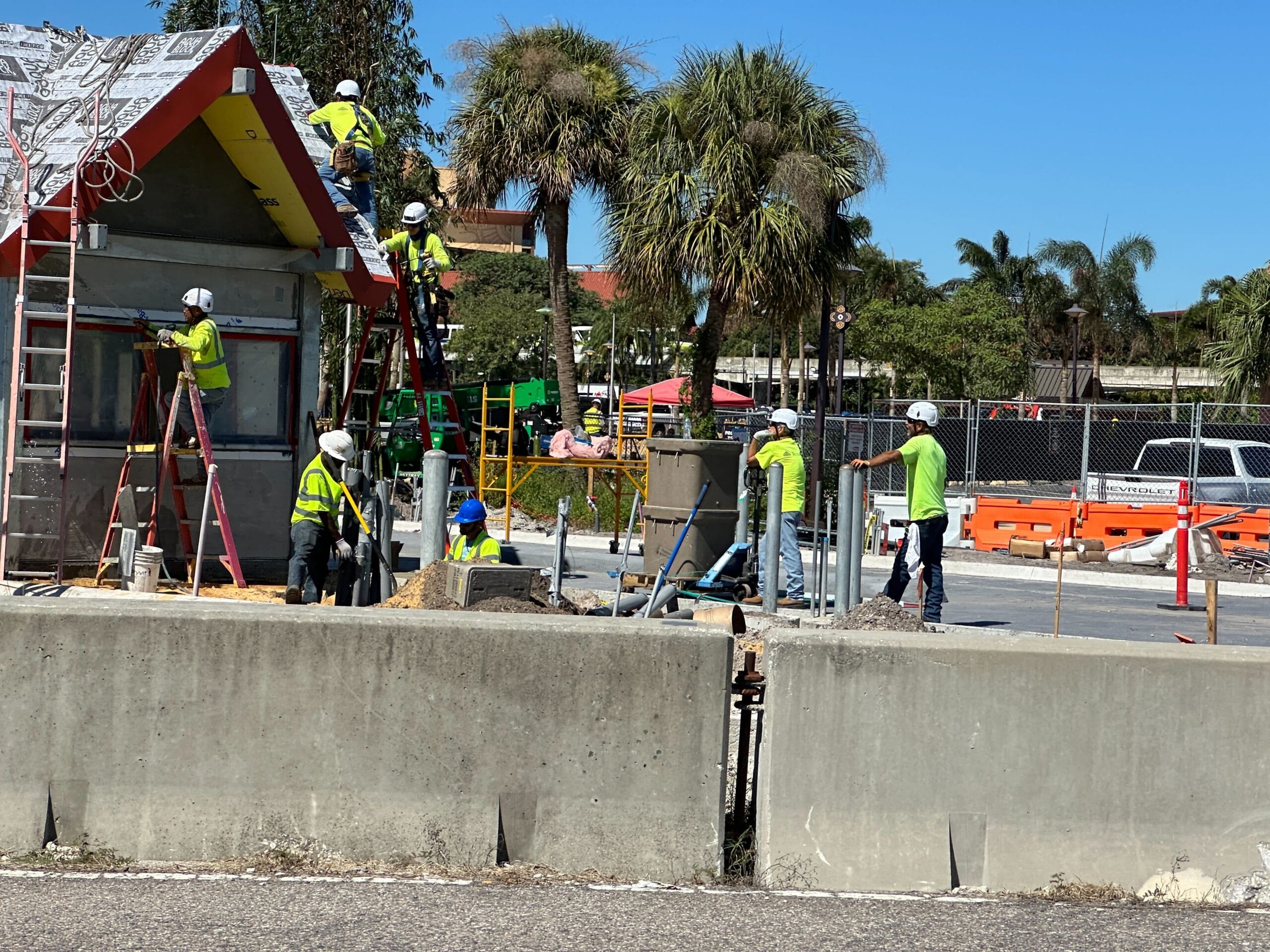 Construction workers in safety gear install doors at a palm-lined building site under blue skies, reminiscent of Disney park entrances.