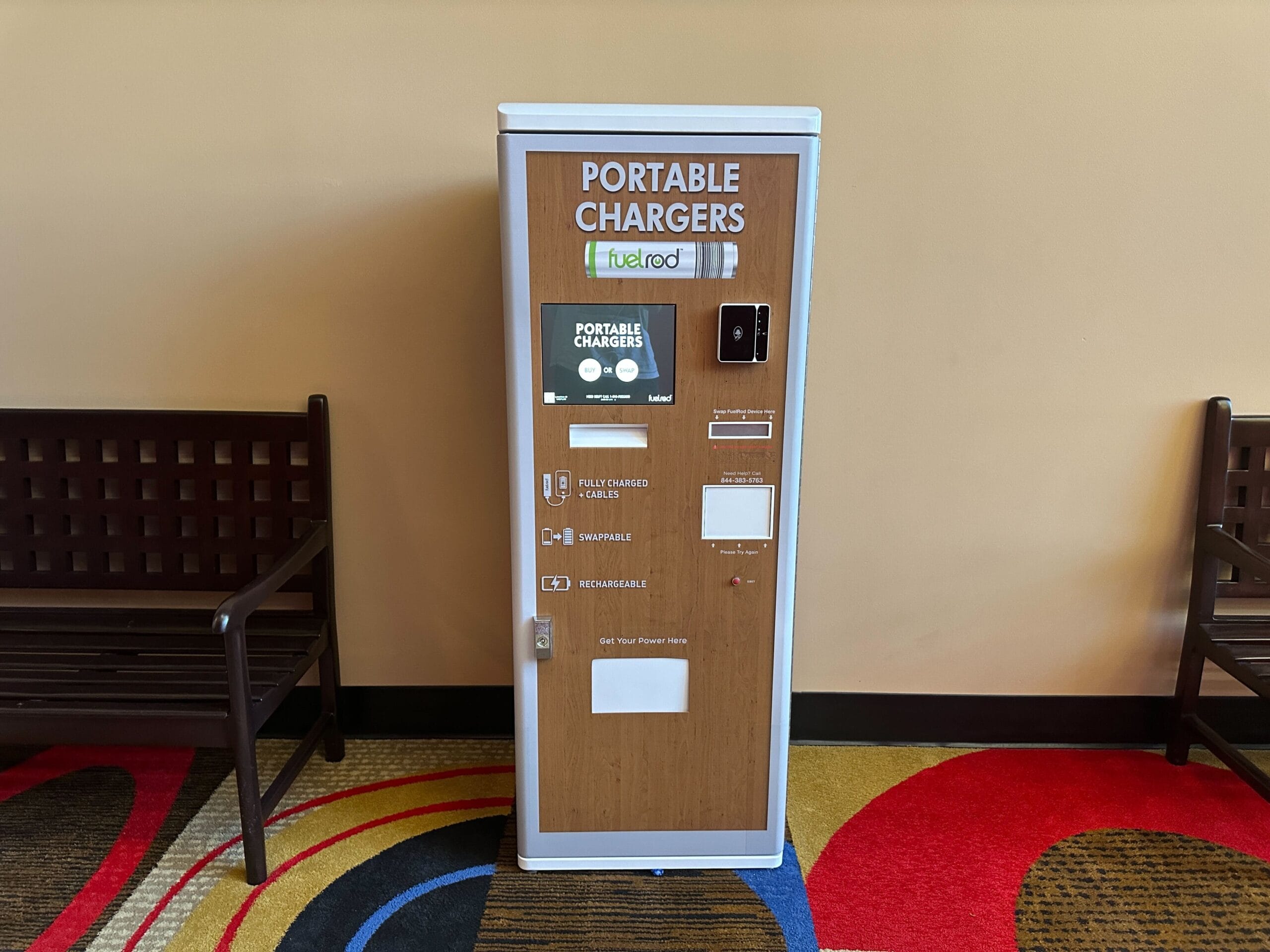 A portable chargers kiosk between two benches sits on a carpet with colorful patterns, like those seen in Disney park lounges.