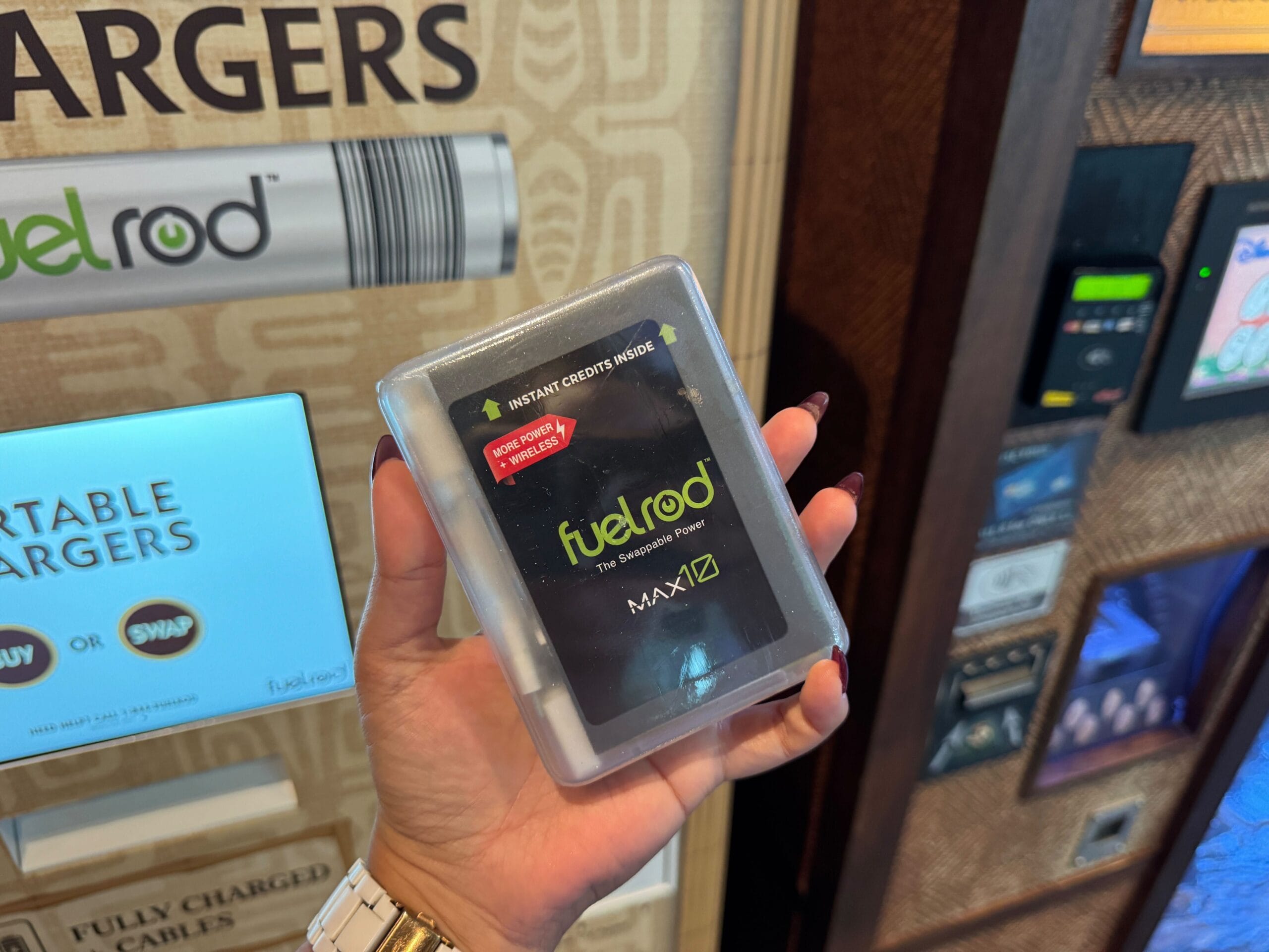 A hand holds a FuelRod charger in front of a FuelRod vending machine at Walt Disney World, home to Cinderella Castle.