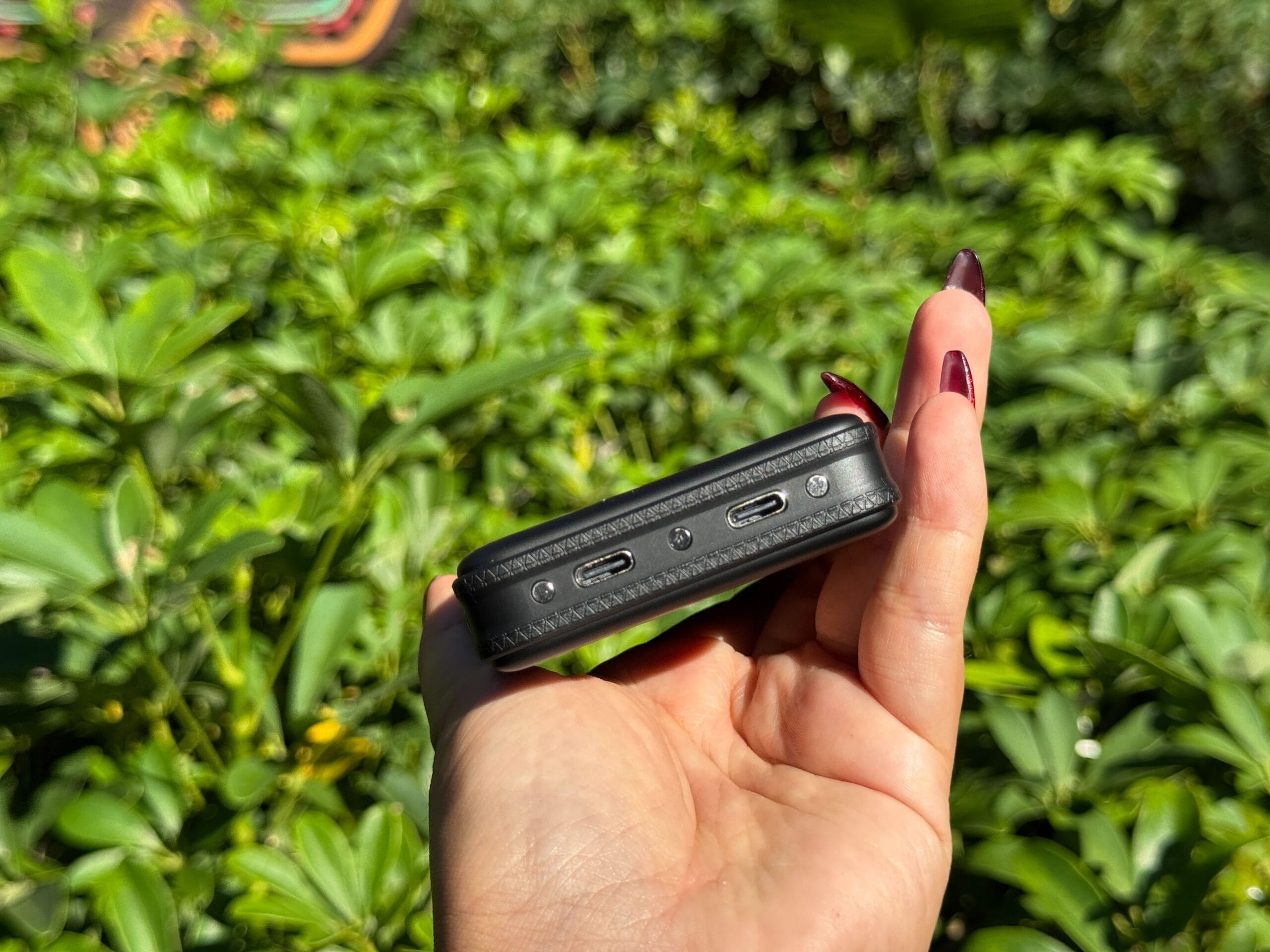 A hand with painted nails holds a MAX10 Option device with two ports, set against lush greenery like that around Magic Kingdom.