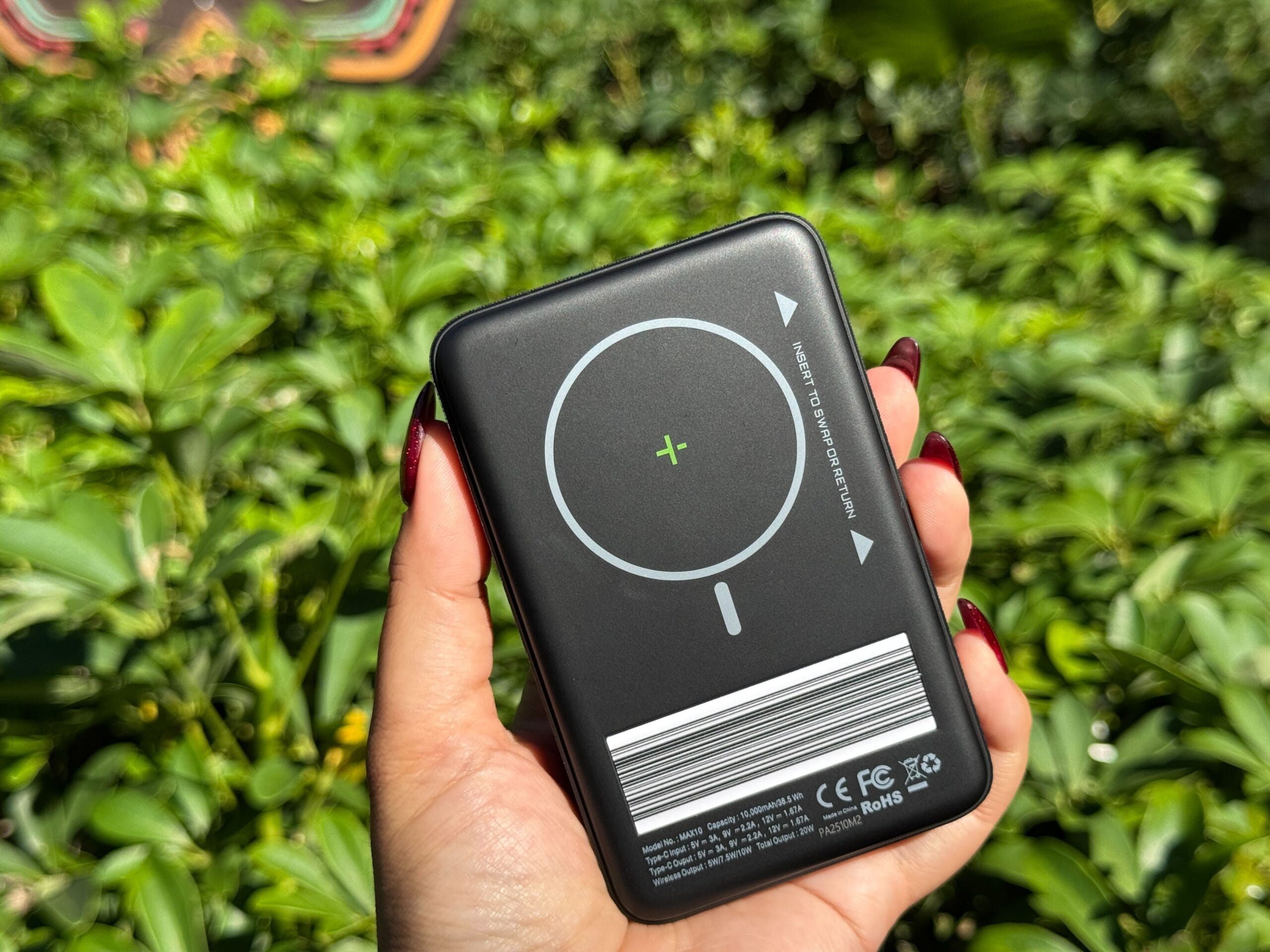 A hand with red nails holds a black FuelRod charger among lush greenery at Walt Disney World, no castle visible for comparison.
