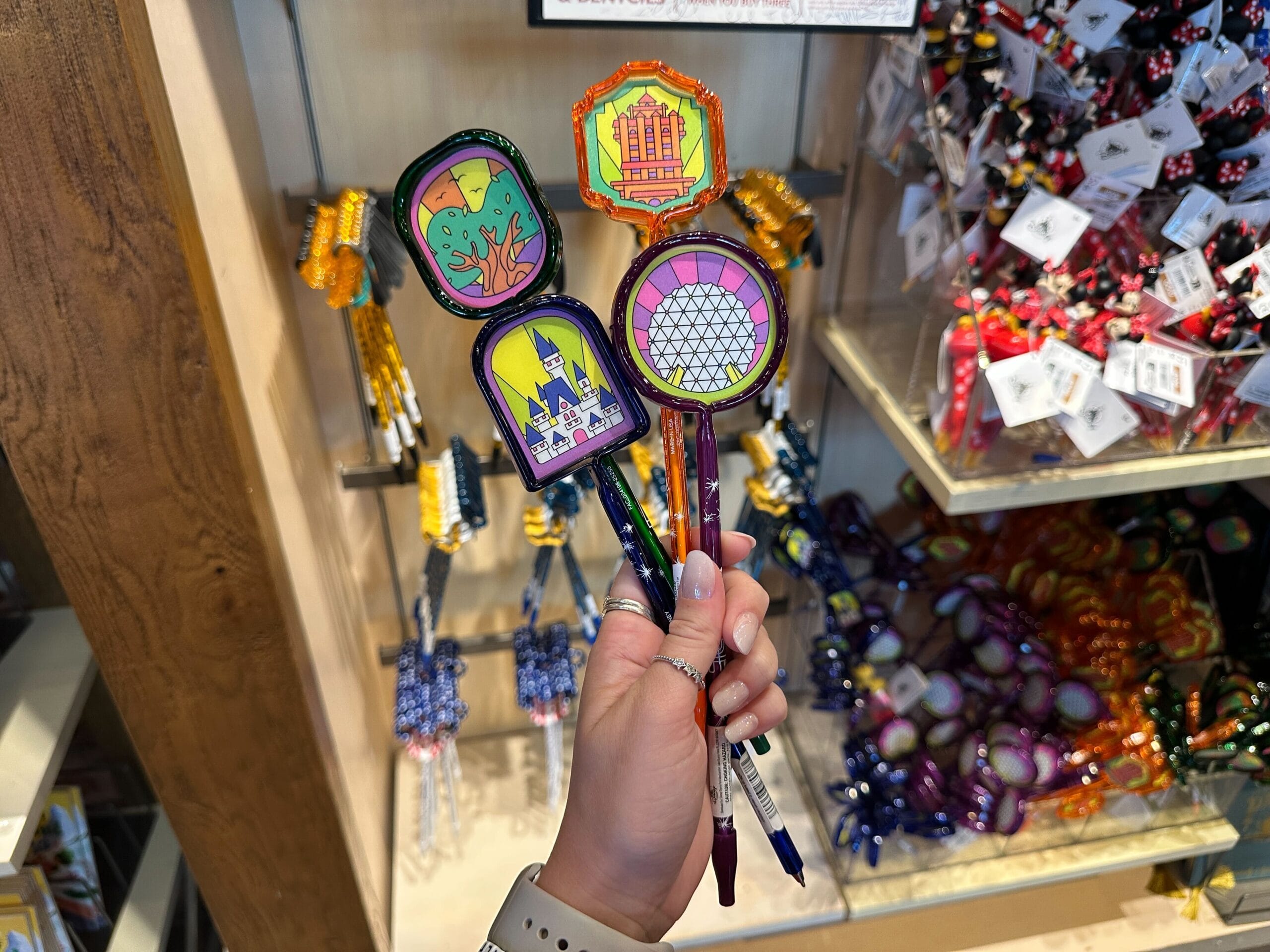 A hand displays Disney World pens with park icons in front of souvenir shelves—no castle shown, but classic Disney theming is evident.