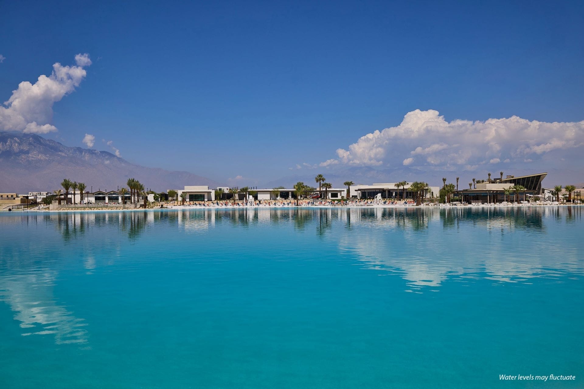 A sparkling blue pool with sleek modern buildings and palm trees, evoking a Disney resort vibe, beneath a bright, cloud-dotted sky.
