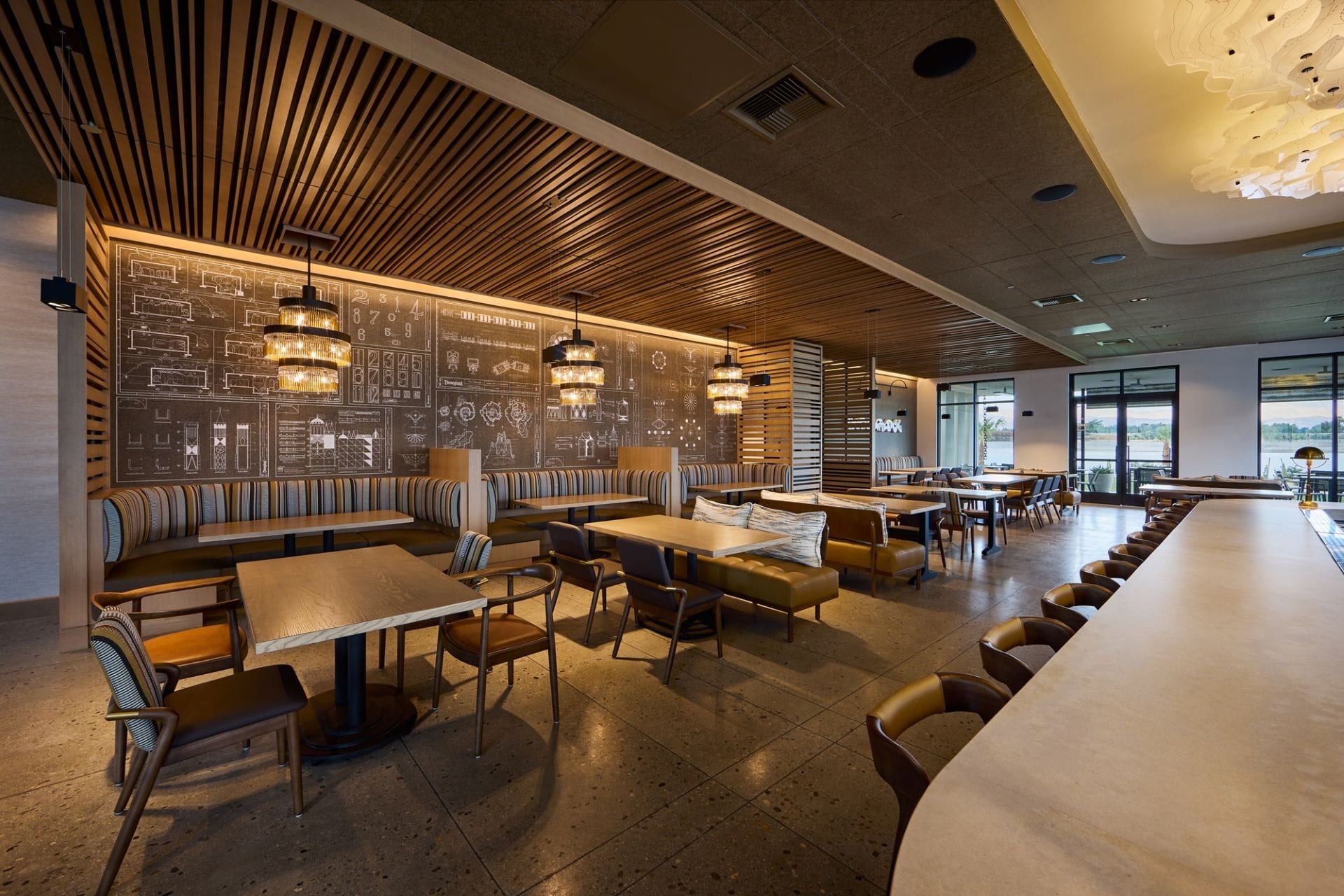 Modern restaurant interior at a Disney park, with wood accents, booths, tables, pendant lights, and large windows facing the grounds.