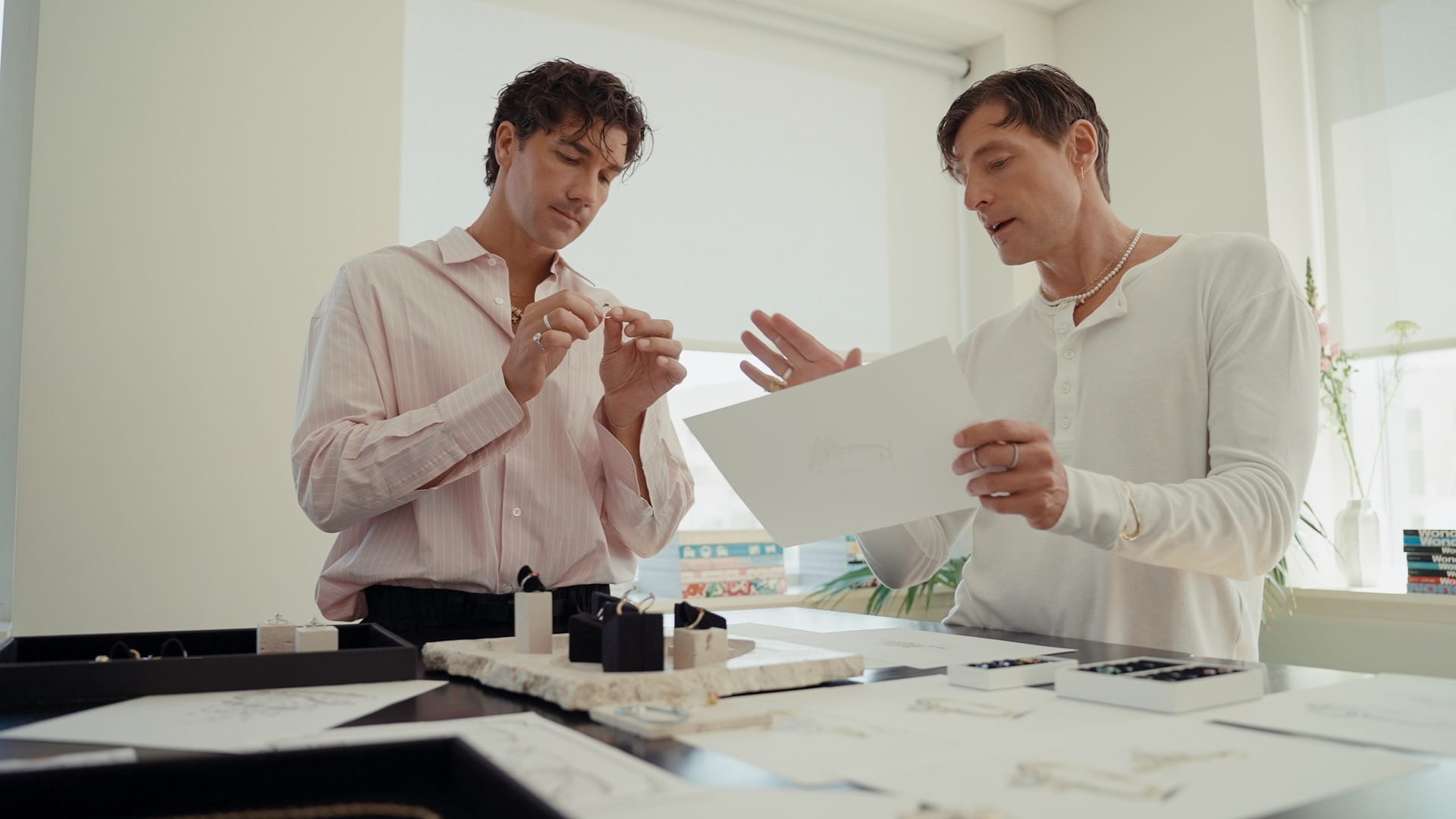 Two men discuss Pandora jewelry designs at a table with sketches and tools, in a bright office reminiscent of Disney park boutiques.
