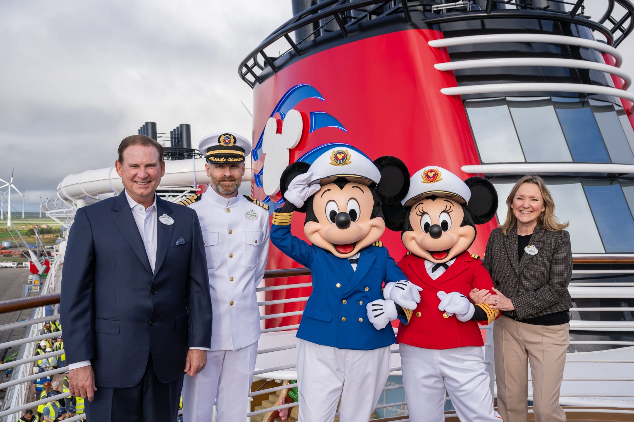 Four adults and a ship captain pose with Mickey and Minnie in cruise captain outfits on a Disney cruise ship deck. No castle visible.