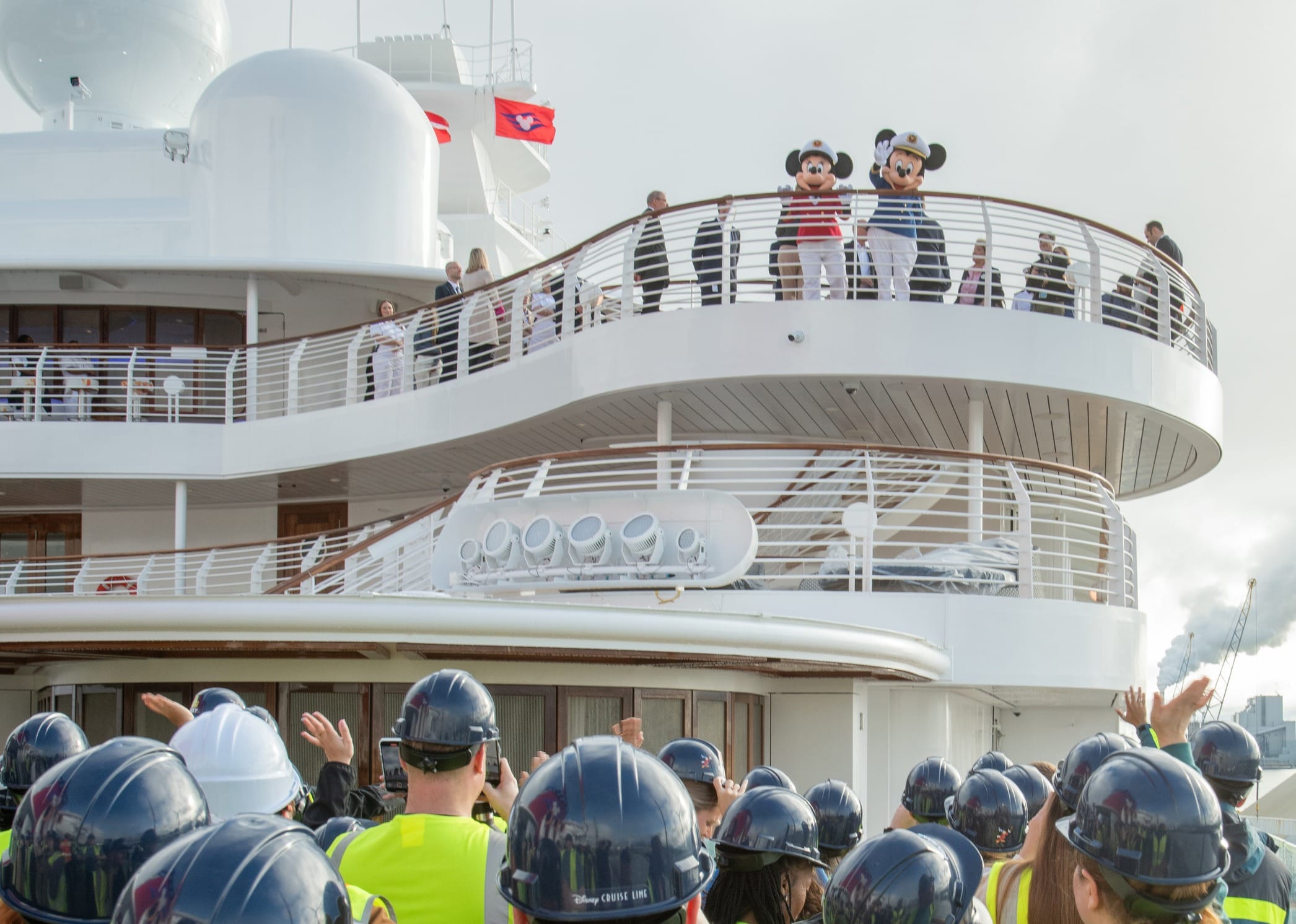 Crew in hard hats wave to Mickey and Minnie on the Disney Destiny cruise ship’s balcony, celebrating the ship’s arrival.