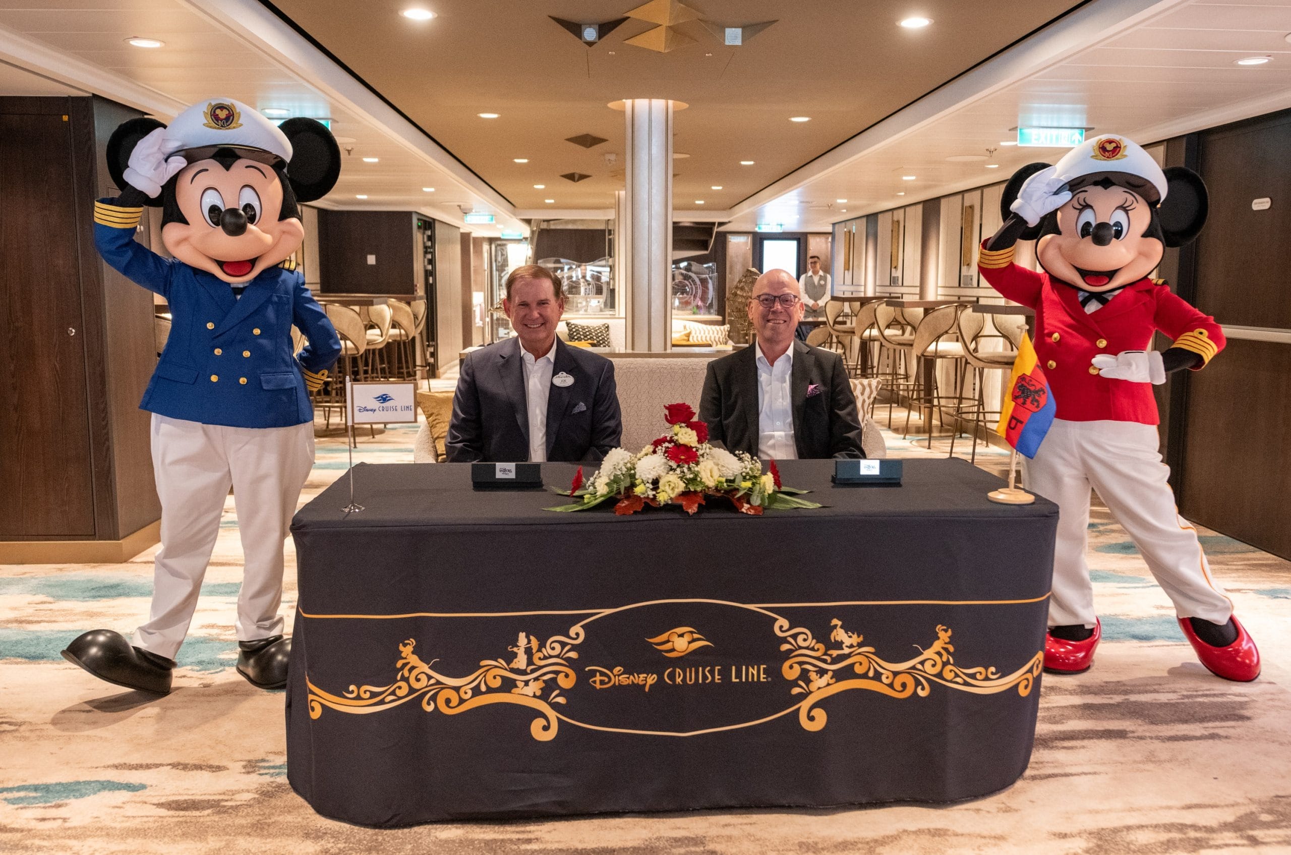 Two men sit at a Disney Cruise Line table with captain Mickey and Minnie beside them, reminiscent of Disney Parks' character meet-ups.