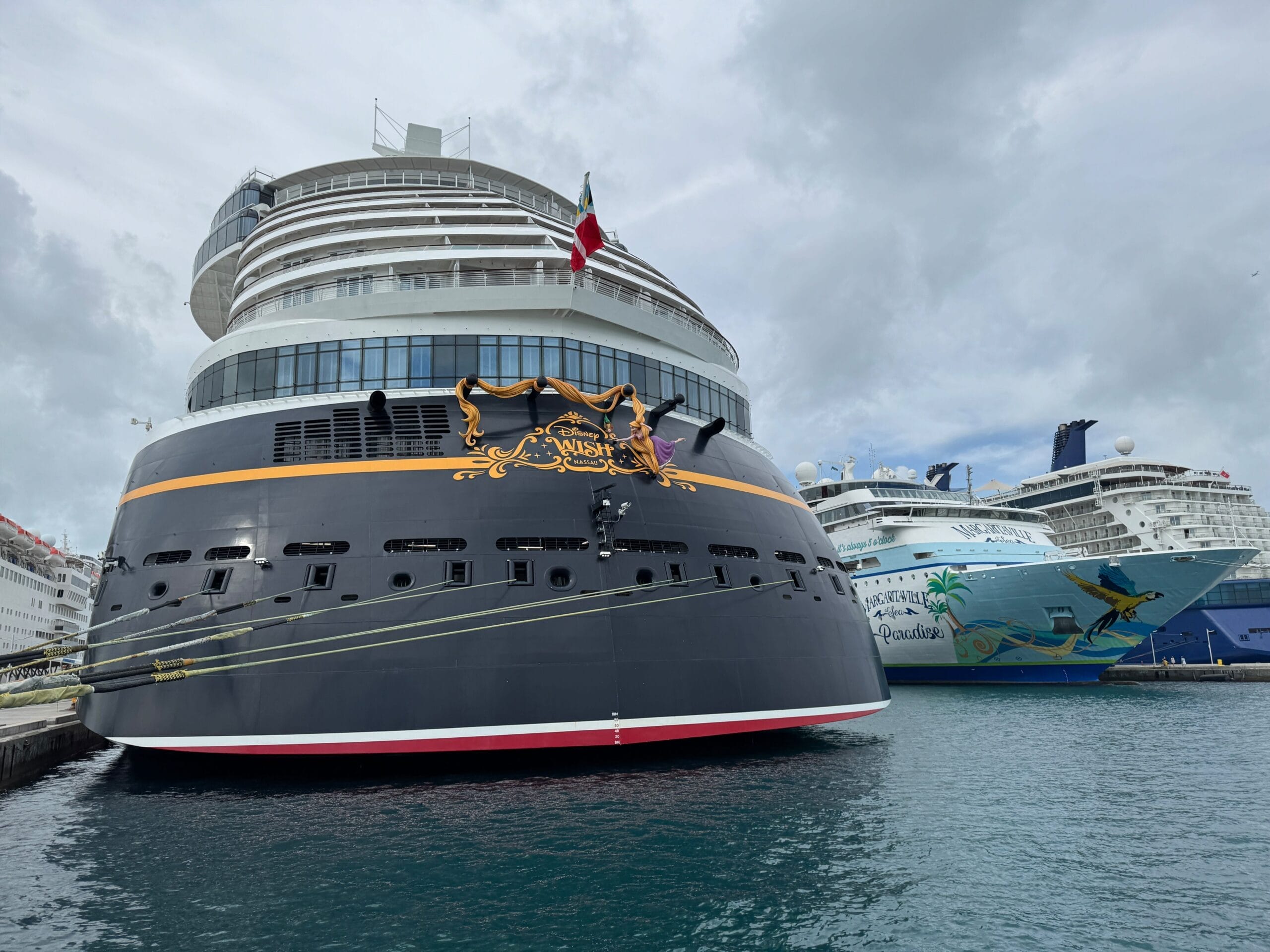 Two large cruise ships, one sporting iconic Disney Cruise Line details, are docked at a port beneath cloudy skies.