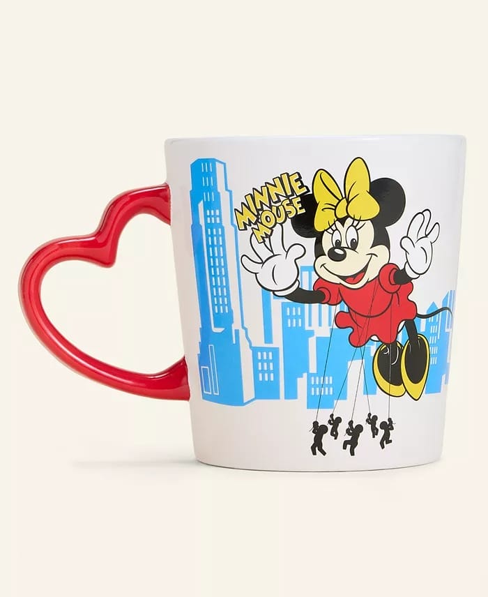 A white mug with a red heart handle, 2025 holiday edition, shows Minnie Mouse as a marionette above blue buildings, no castle shown.