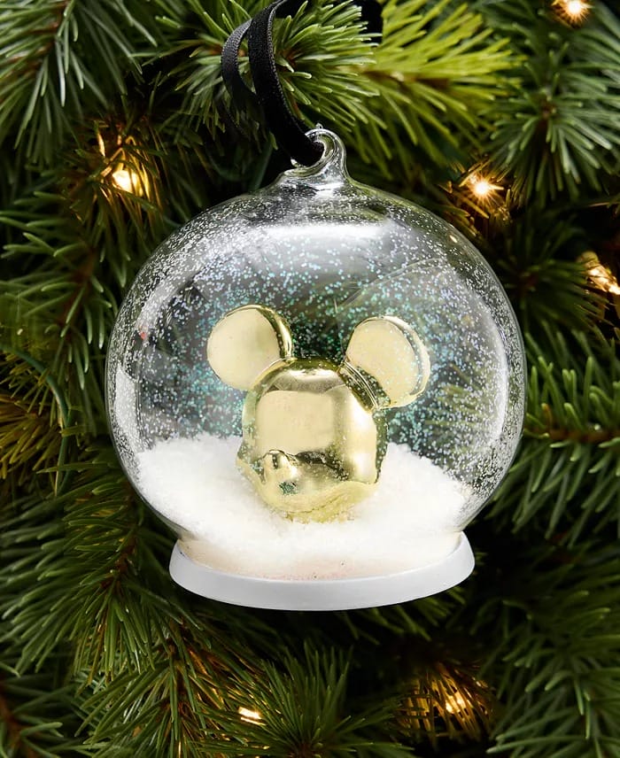 A gold mouse-head glass ornament from the Disney 2025 Holiday Collection hangs on a sparkling tree, reminiscent of park decor.