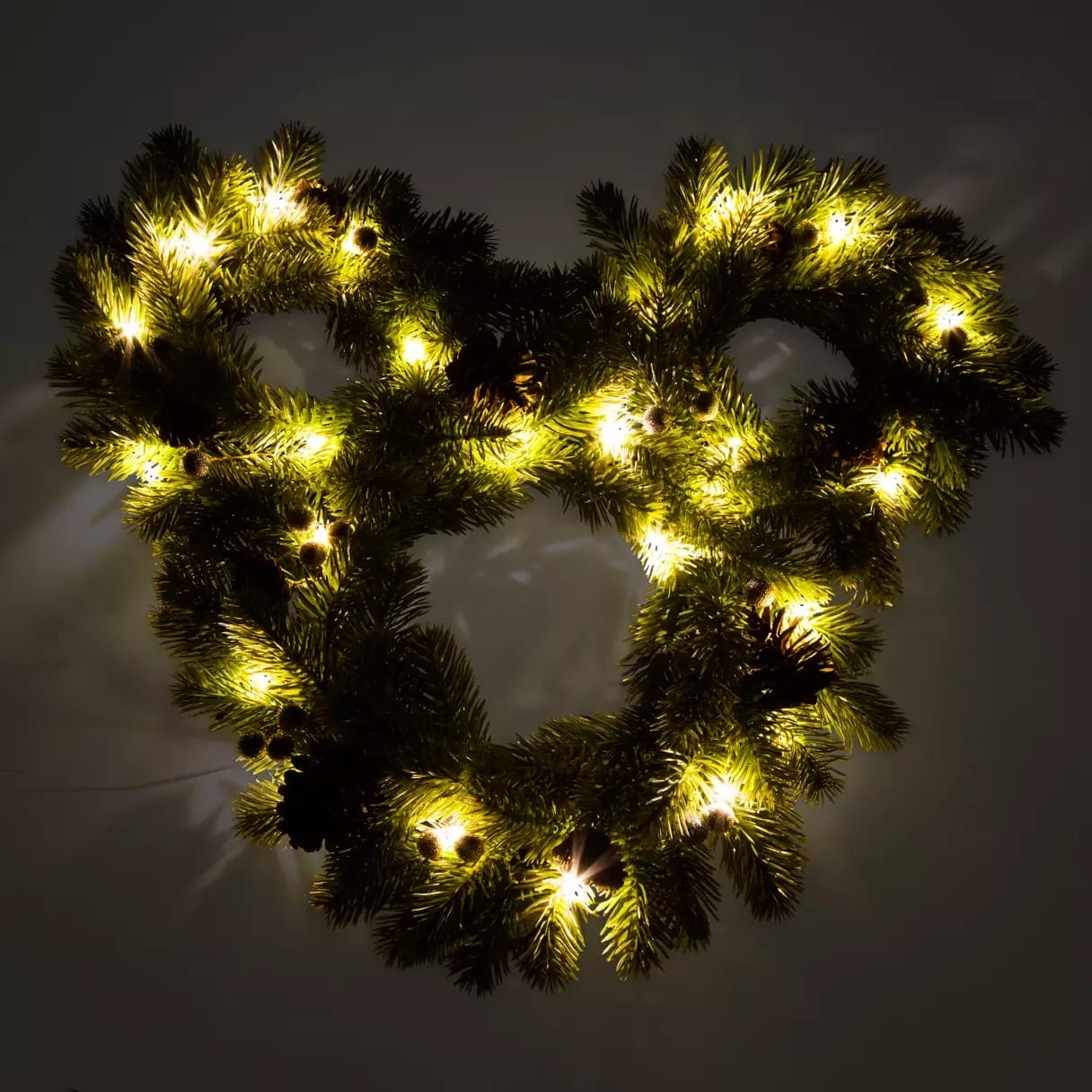 A Mickey Mouse-shaped Christmas wreath with pine branches and warm white lights glows on a wall.