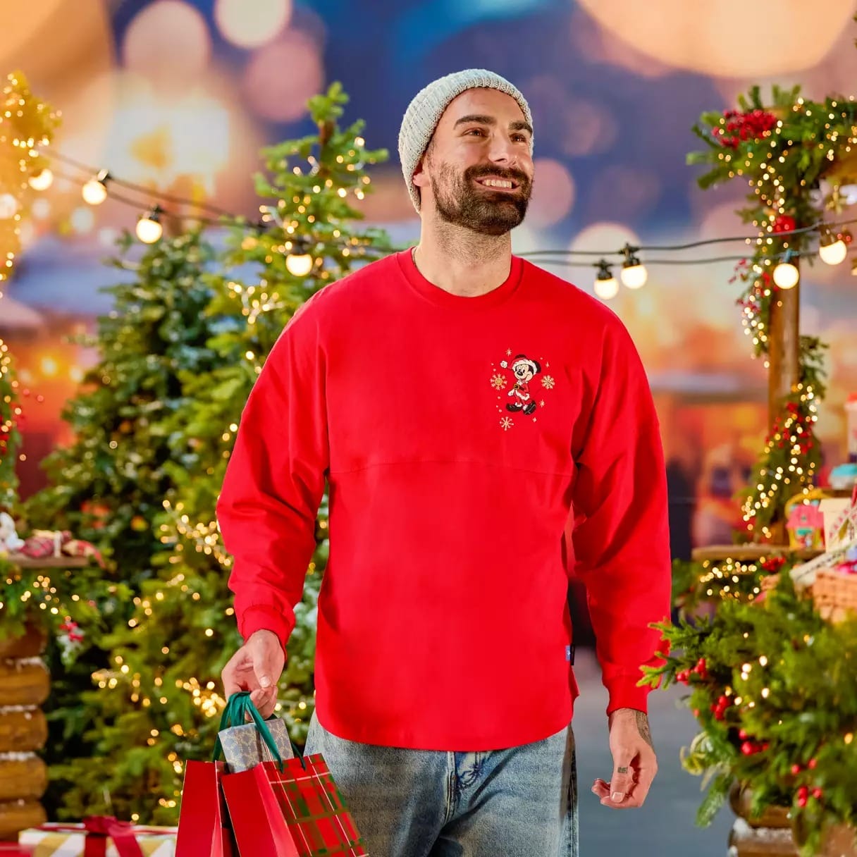 Man in red sweater and beanie holding gift bags, standing among decorated Christmas trees with festive lights.